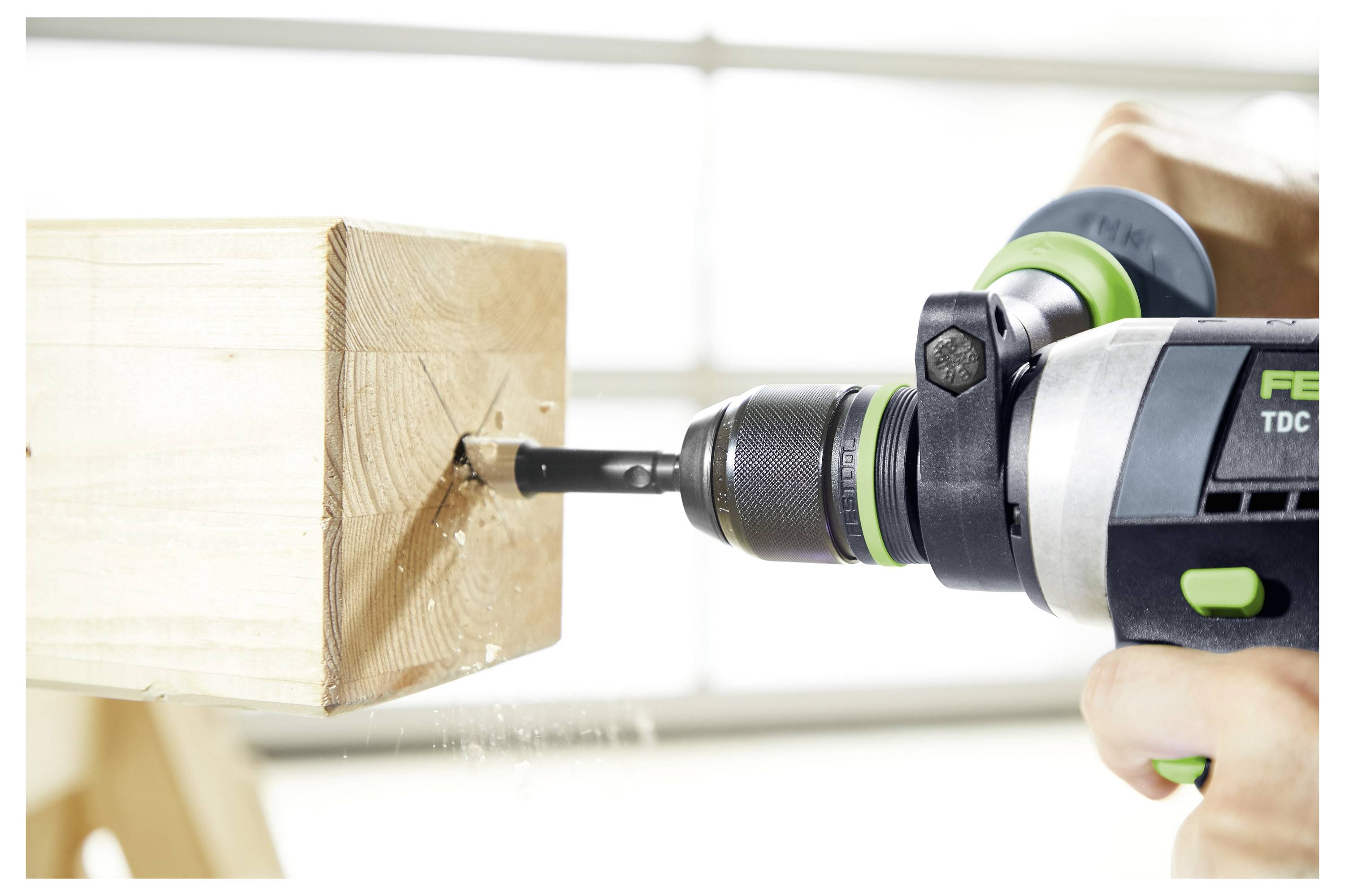 Eine Person bedient eine Elektro-Gehrungssäge, um ein Holzbrett im Freien zu zuschneiden. Im Hintergrund ist ein Holzgebäude zu sehen.