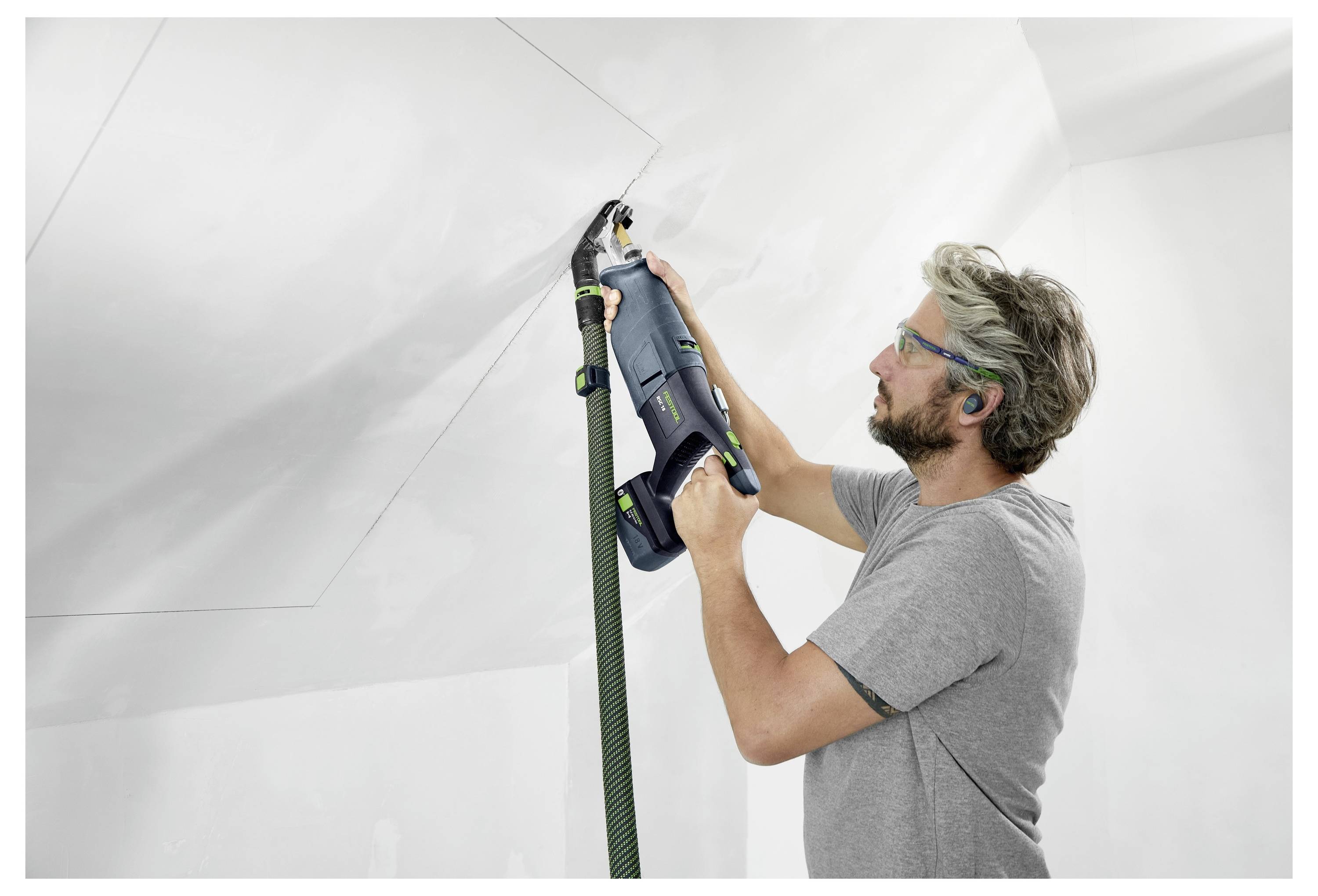 A man wearing safety glasses uses a power tool to sand a ceiling, ensuring a smooth surface. He holds the vacuum hose attached to the tool.
