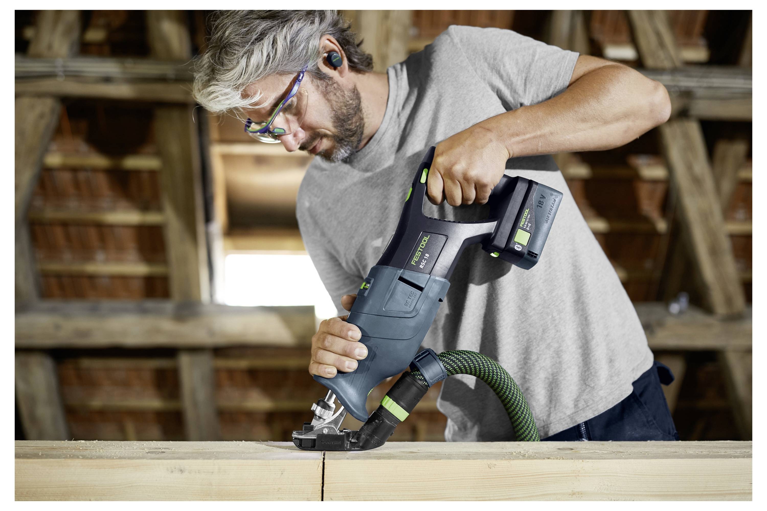 Eine Person trägt eine Schutzbrille und benutzt ein Elektrowerkzeug zum Holzschneiden in einer Werkstätte. Sie konzentriert sich auf die Aufgabe, wobei eine Holzdachkonstruktion im Hintergrund sichtbar ist.