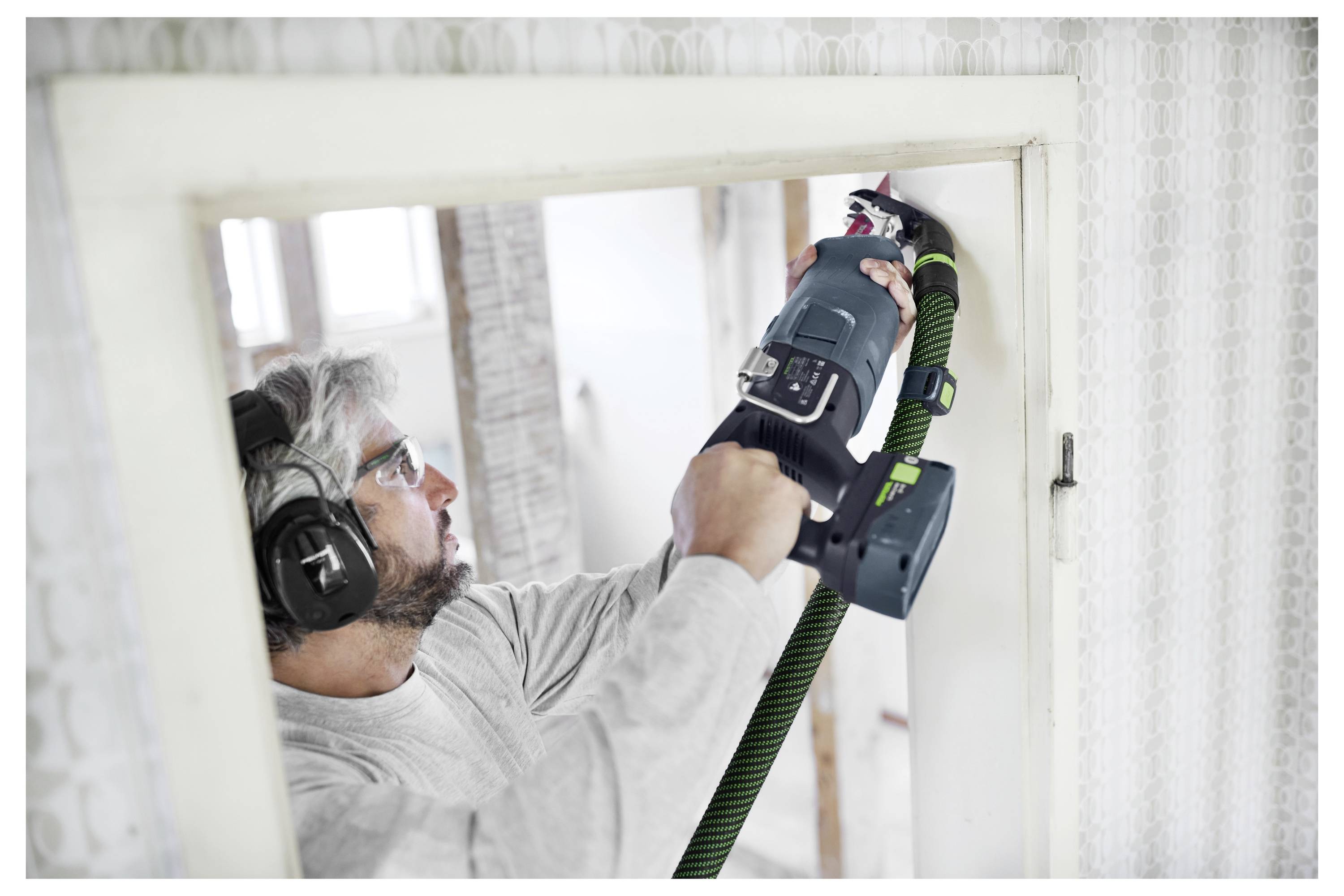 Eine Person, die eine Schutzbrille und Gehörschutz trägt, verwendet ein Elektrowerkzeug, um durch eine Wand in einem hell erleuchteten Raum unter Renovierung zu schneiden.