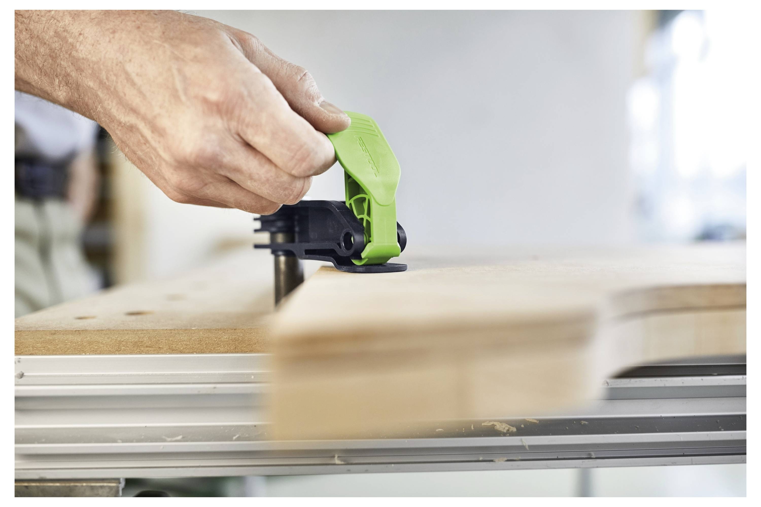 Eine Hand, die ein grünes Werkzeug verwendet, um ein Holzbrett sicher an einer Werkbank festzuspannen, was einen laufenden Holzbearbeitungsprozess veranschaulicht.