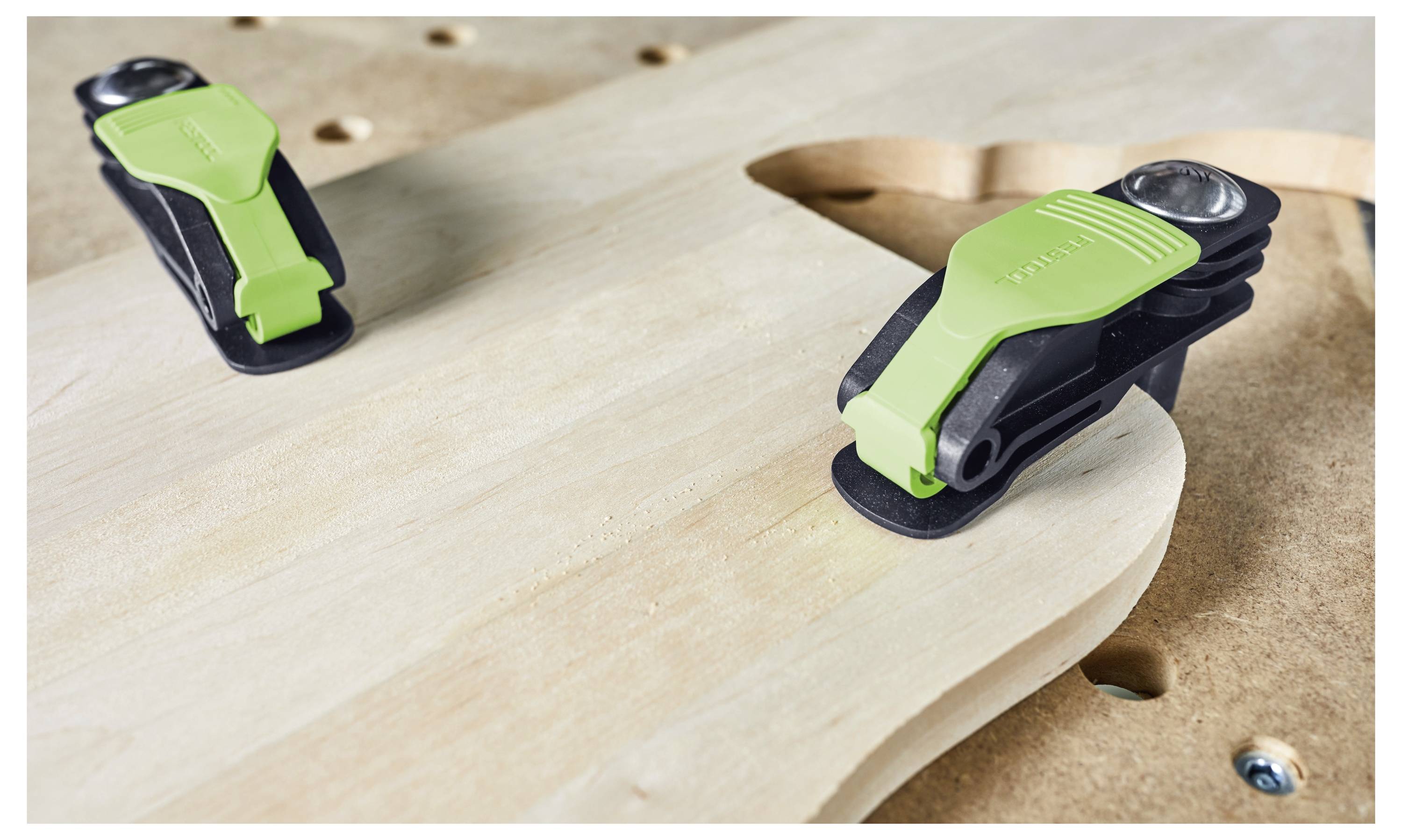 Zwei schwarze und grüne Klemmen befestigen ein Holzbrett sicher an einem Tisch und deuten auf ein laufendes Holzbearbeitungsprojekt hin.