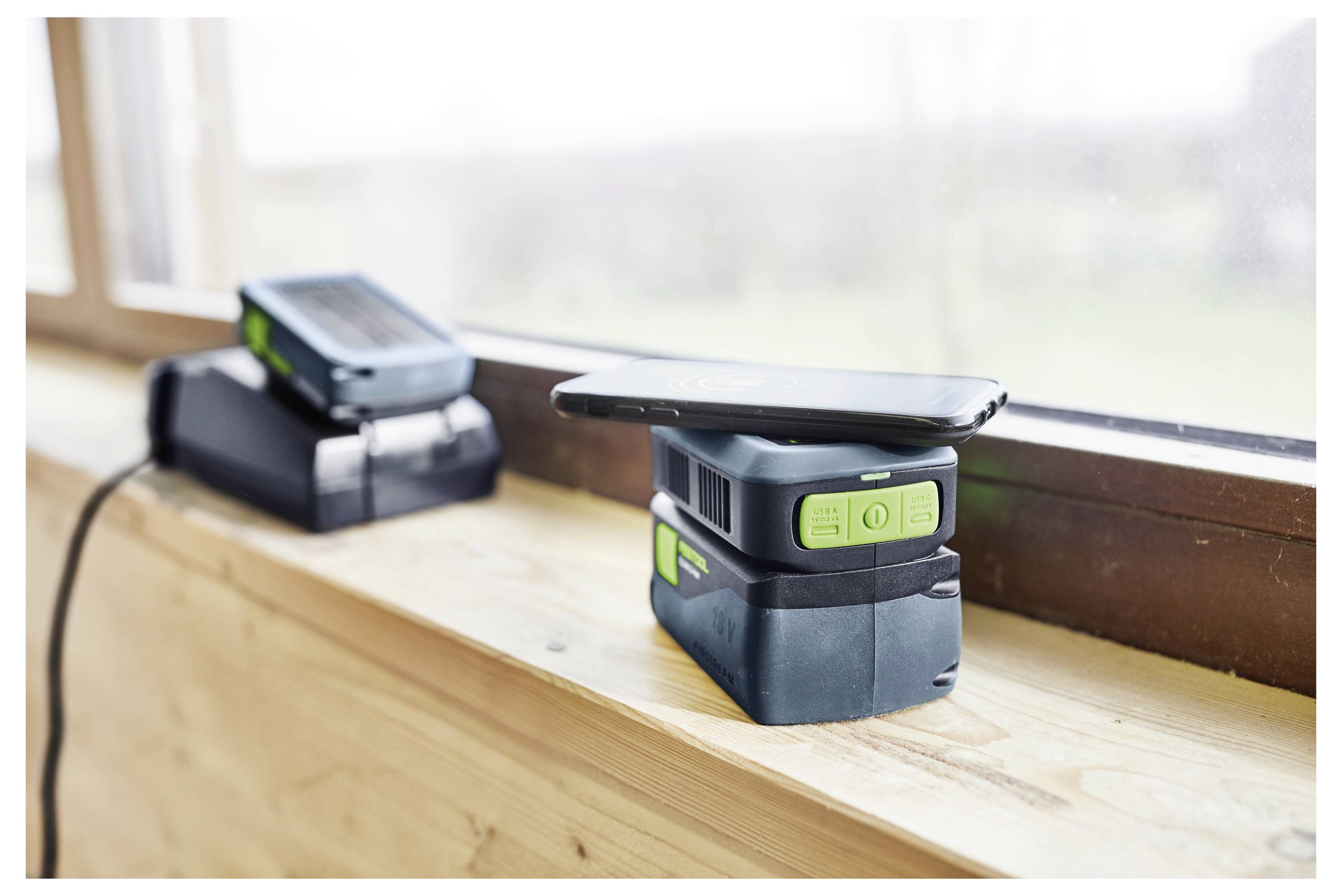 Ein Smartphone wird kabellos auf einer Powerbank aufgeladen, die auf einer hölzernen Fensterbank liegt. Ein weiteres Gerät wird im Hintergrund aufgeladen.