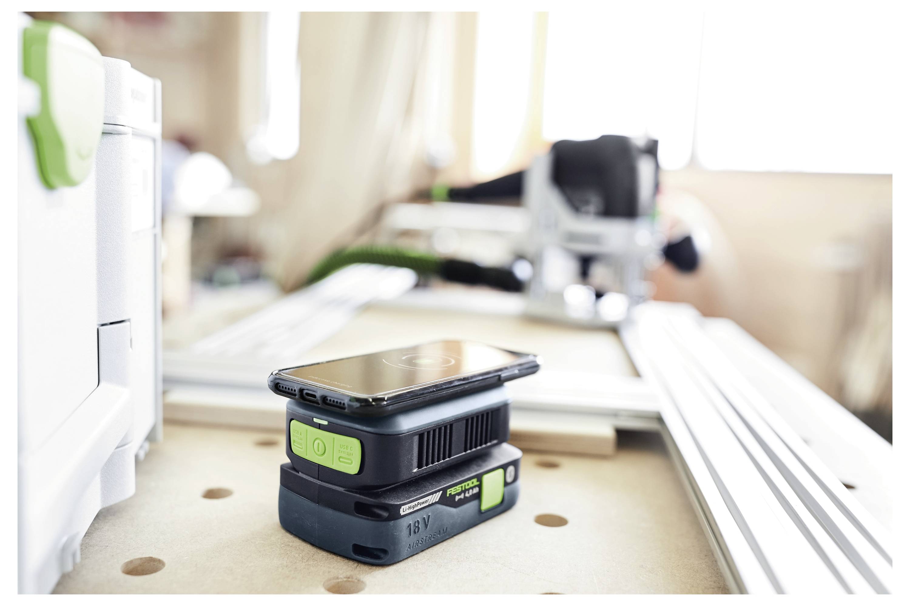 Ein Smartphone ruht auf einer schwarz-grünen Elektrowerkzeug-Batterie auf einer Werkbank, mit unscharfen Werkstattwerkzeugen im Hintergrund.