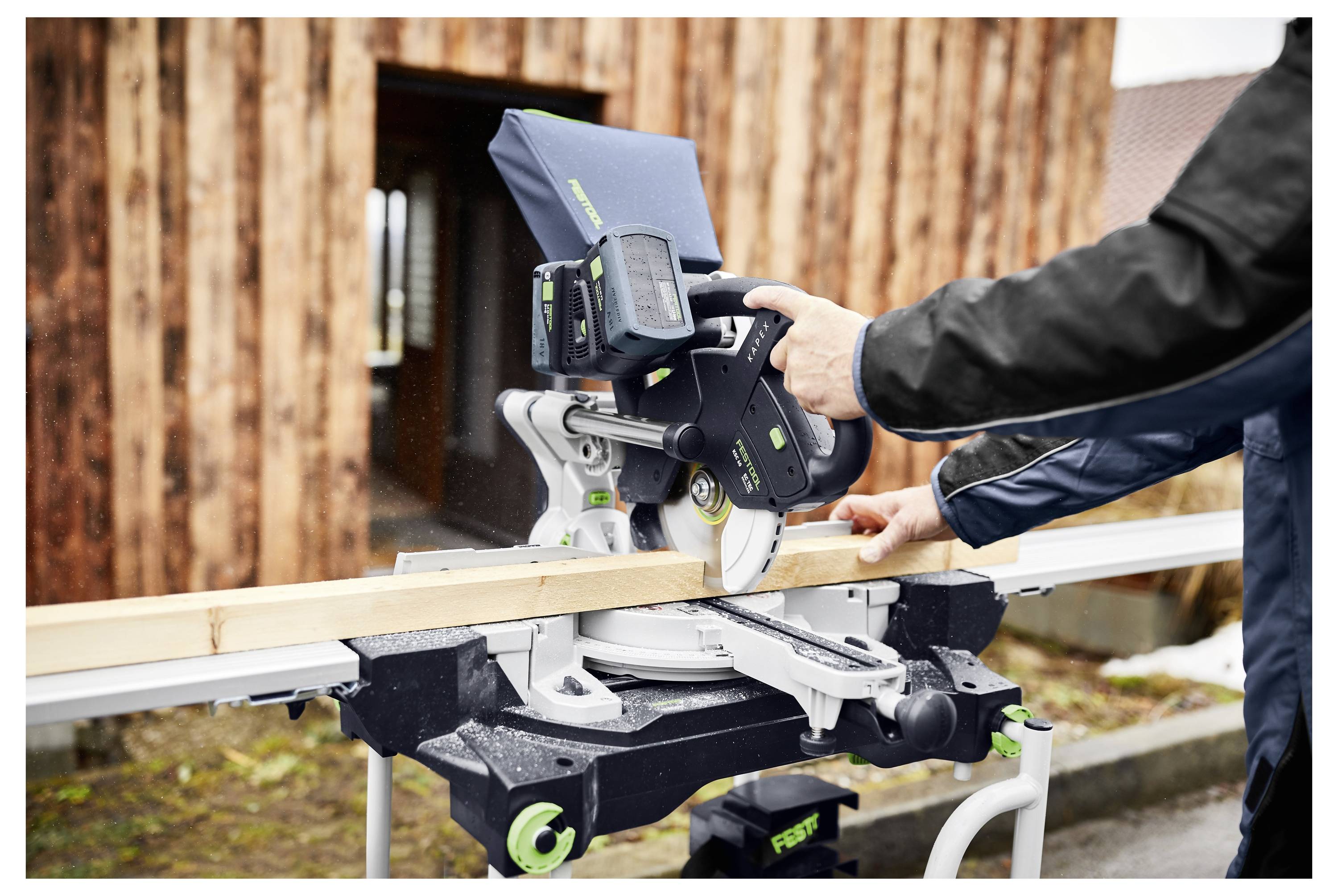 A person operates a power miter saw to cut a wooden plank outdoors. The background features a wooden building.