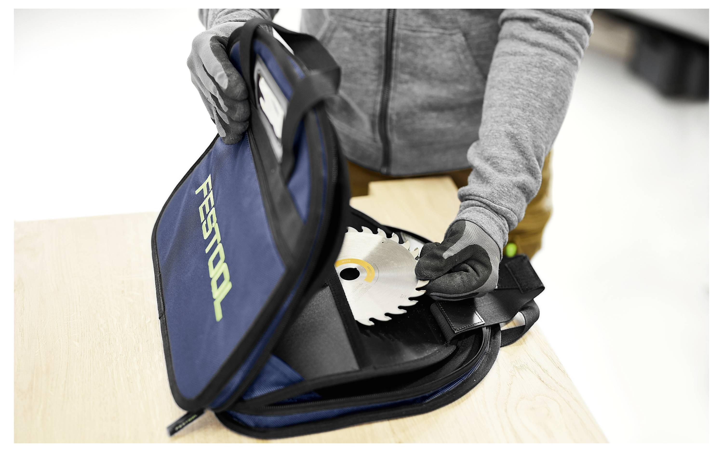 Eine Person mit Handschuhen legt ein kreisrundes Sägeblatt in einen blauen Festool-Beutel auf einem Holztisch, was auf sichere Beförderung oder Lagerung hindeutet.