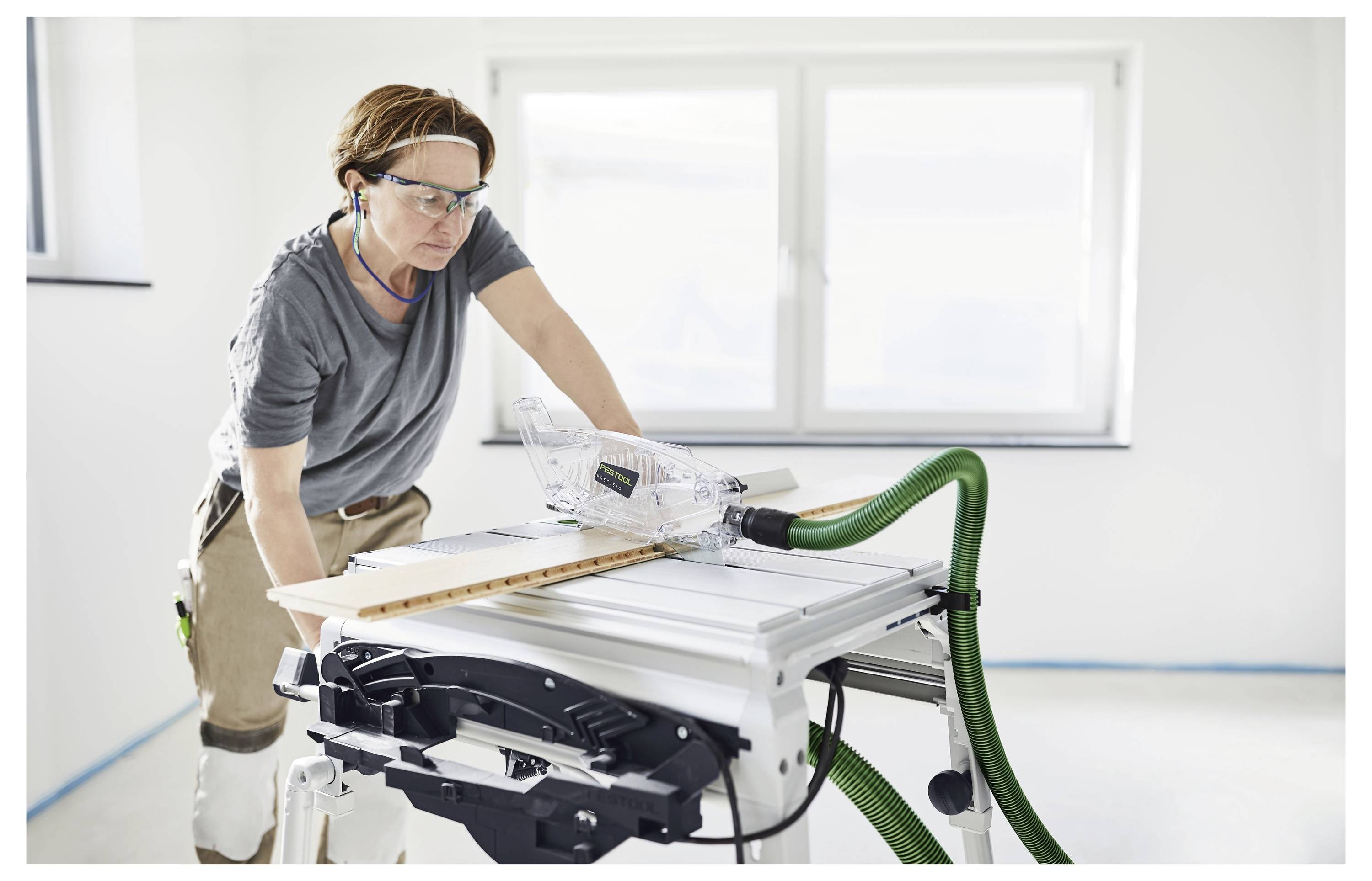 Person, die eine Tischkreissäge nutzt, um Holz in Innenräumen zu schneiden, trägt eine Schutzbrille und Kopfhörer, mit konzentriertem Gesichtsausdruck. Heller, minimalistischer Raum.