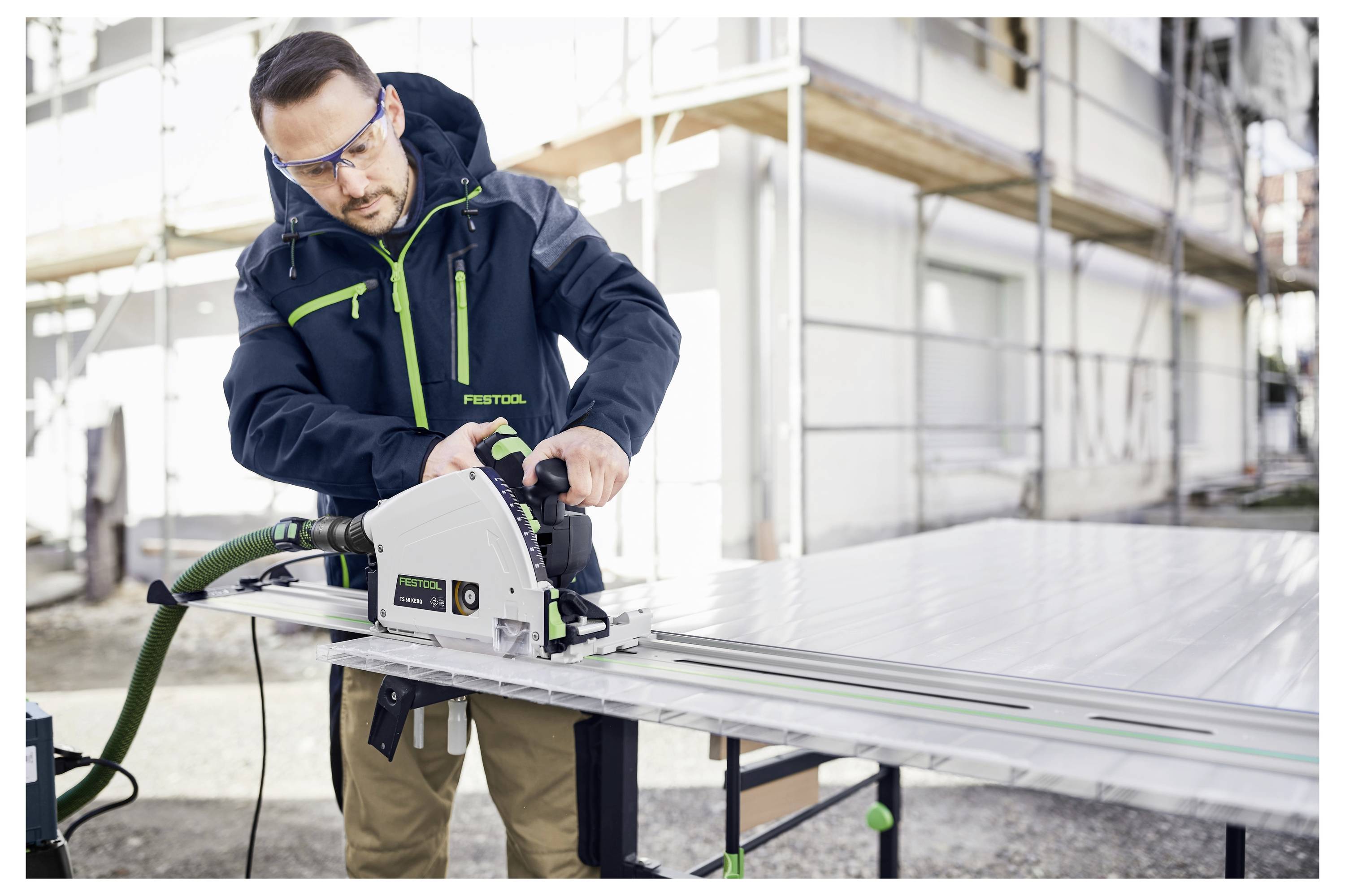 Eine Person in Schutzbrille benutzt eine Festool Tauchschnitt-Schiene auf einem großen weißen Panel im Freien auf einer Baustelle.
