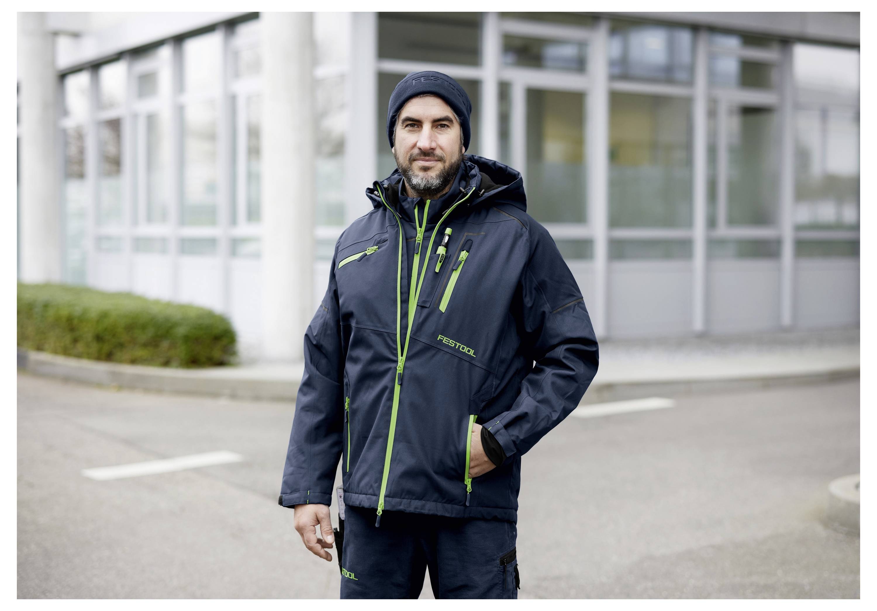 Eine Person in einer dunklen Winterjacke und einem Hut steht vor einem modernen Gebäude und blickt in die Kamera.