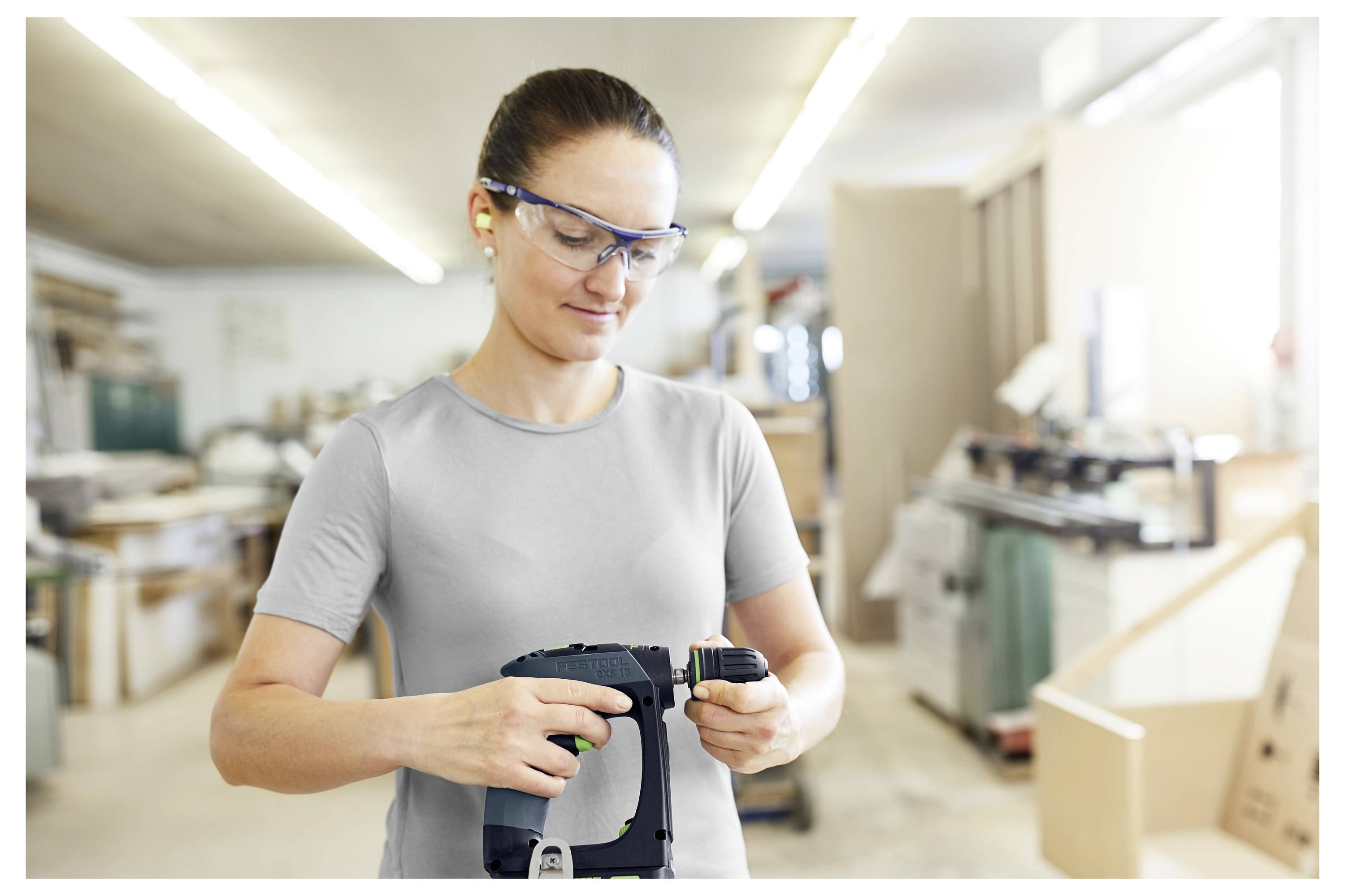 Eine Person mit Schutzbrille arbeitet in einer Werkstatt und bedient ein Elektrowerkzeug mit großer Konzentration. Der Hintergrund ist verschwommen und zeigt verschiedene Geräte.