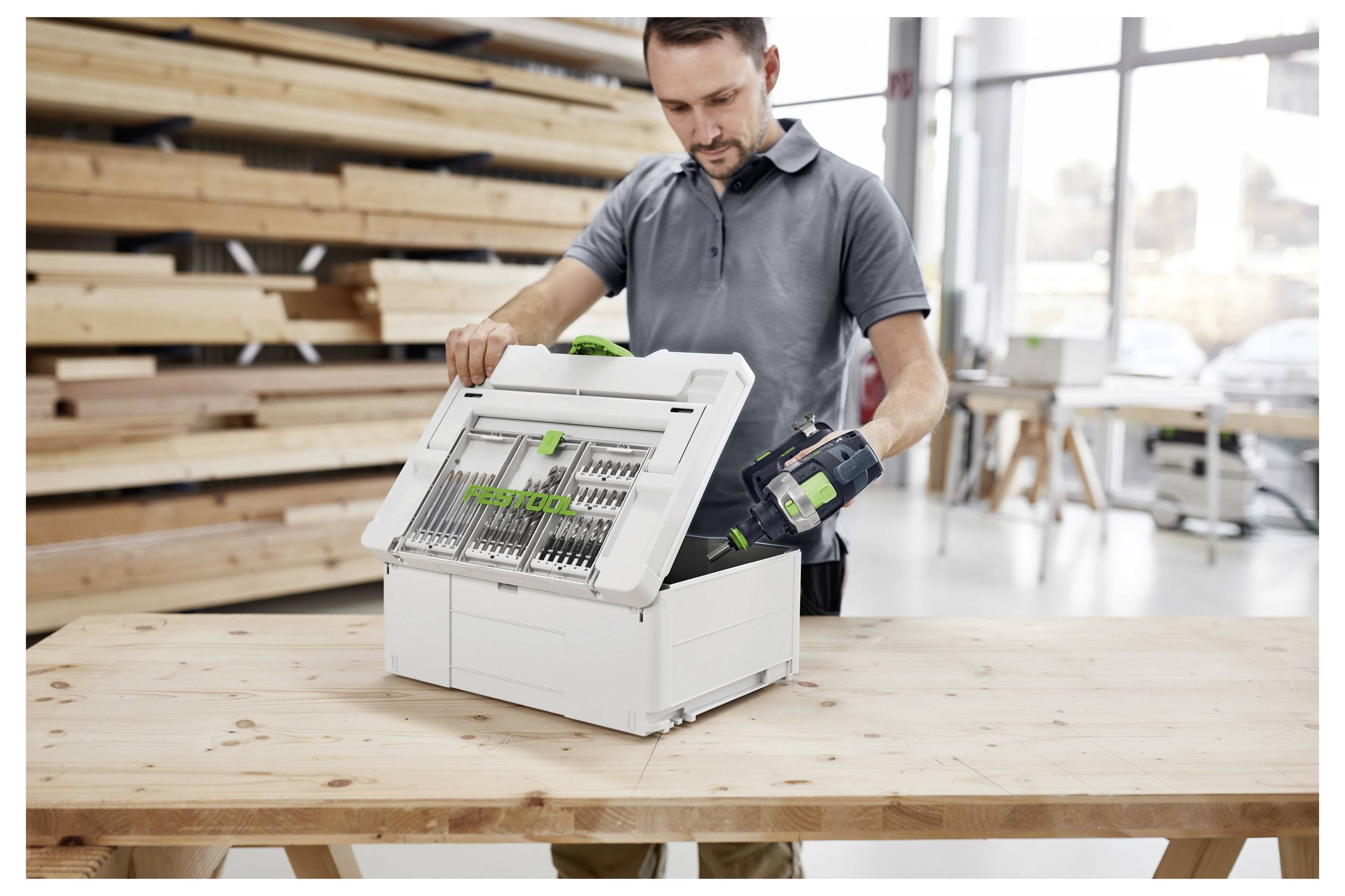 Eine Person in einer Werkstatt öffnet eine weiße Box mit organisierten Werkzeugen. Holzbretter und große Fenster sind im Hintergrund zu sehen.