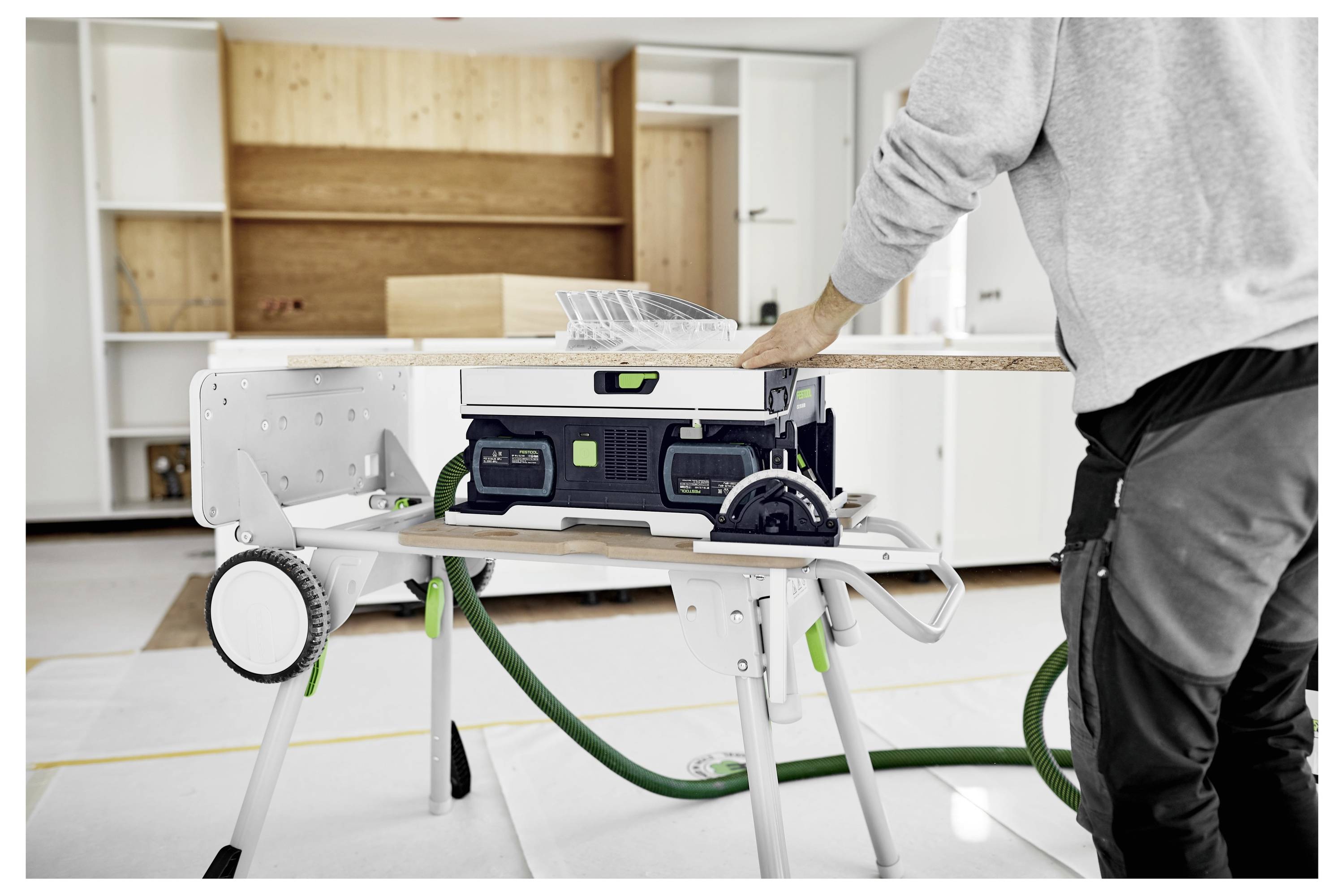 Eine Person in einer Werkstattumgebung schneidet ein Stück Holz auf einer kompakten, tragbaren Tischkreissäge, umgeben von moderner Schrankausrüstung und Werkzeugen.