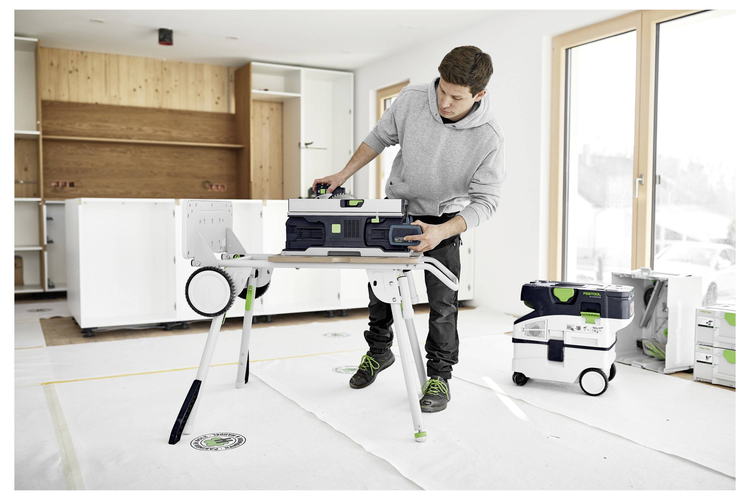 Eine Person in einem grauen Hoodie bedient ein Werkzeug auf einem Ständer in einer teilweise eingebauten Küche, wobei Schränke und Ausrüstung im Hintergrund sichtbar sind.