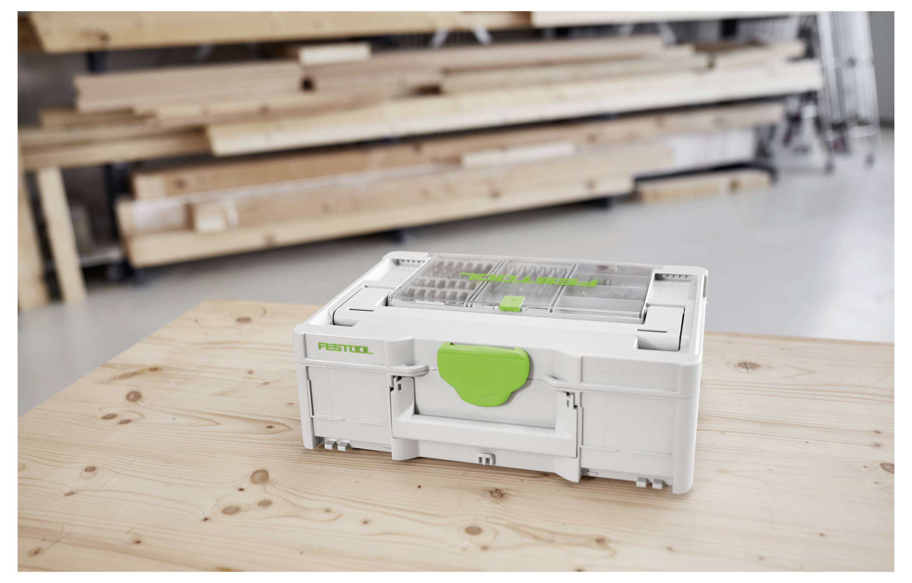 Eine weiße und grüne Werkzeugkiste sitzt auf einem Holztisch in einer Werkstatt, umgeben von unscharfen Holzbrettern im Hintergrund.