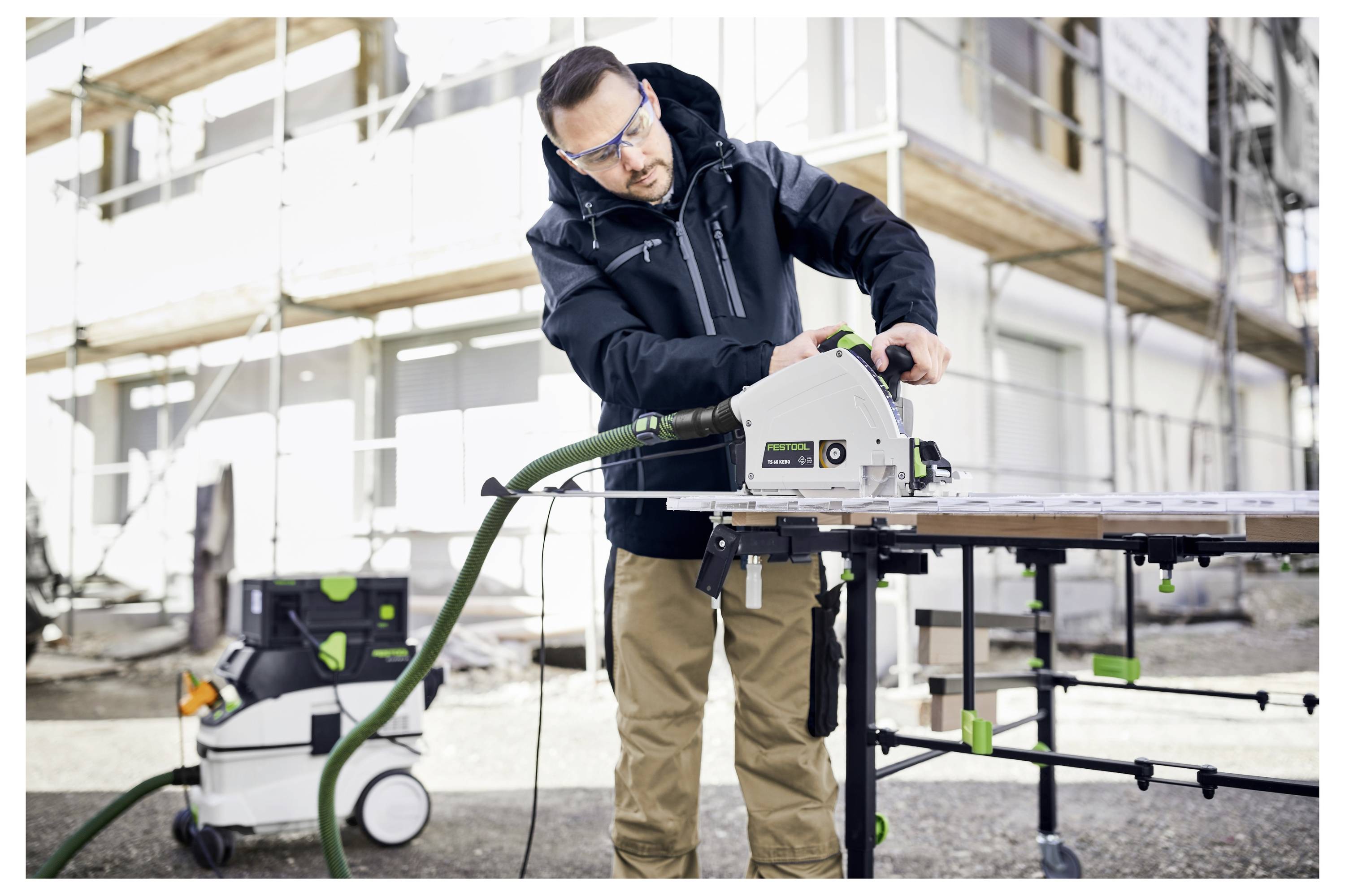 Eine Person mit Schutzbrille bedient eine Kreissäge auf einer Werkbank auf einer Baustelle, mit Gerüsten im Hintergrund.