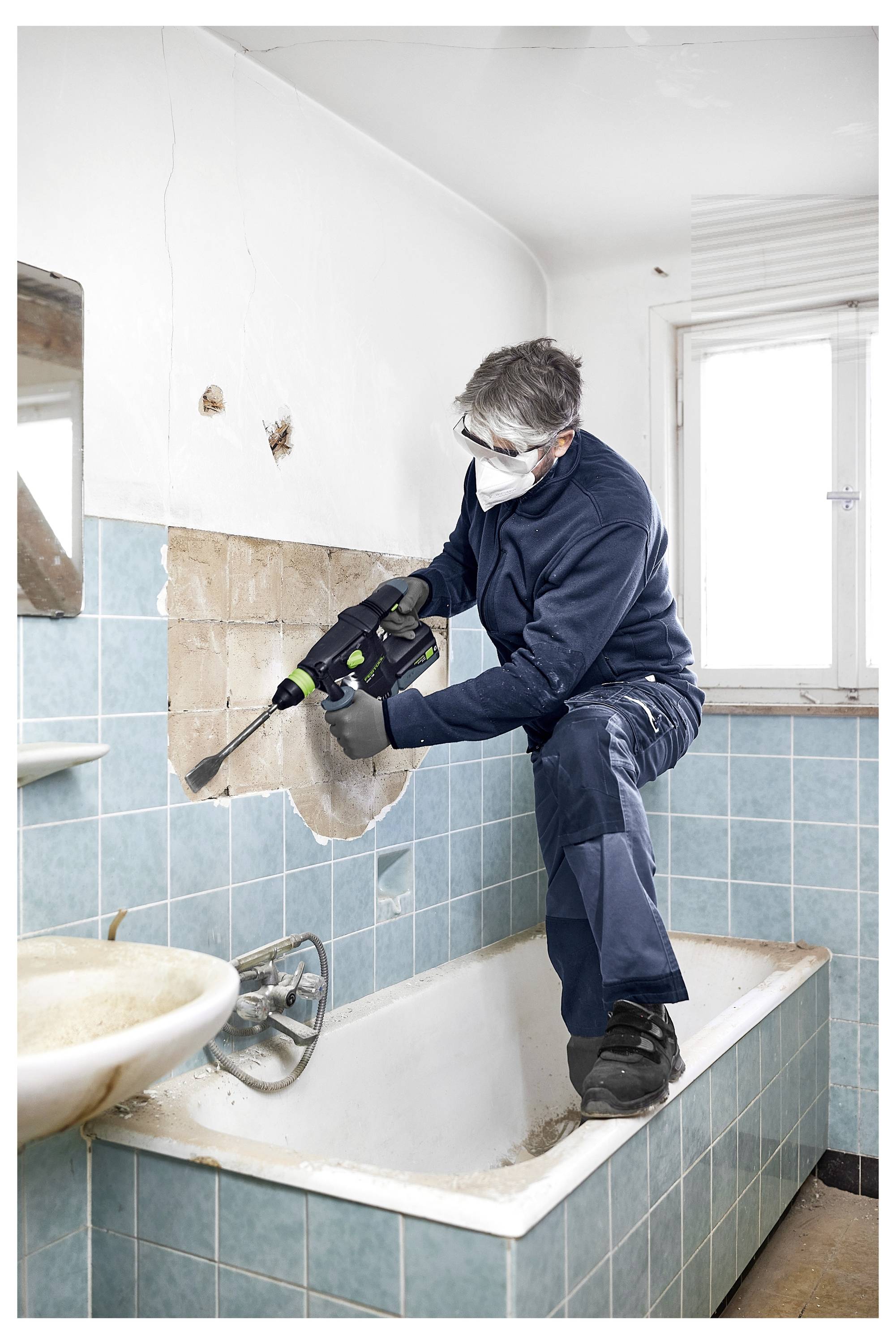 Eine Person in Schutzausrüstung verwendet ein Elektrowerkzeug, um Fliesen von einer Badezimmerwand zu entfernen, während sie in einer Badewanne steht.