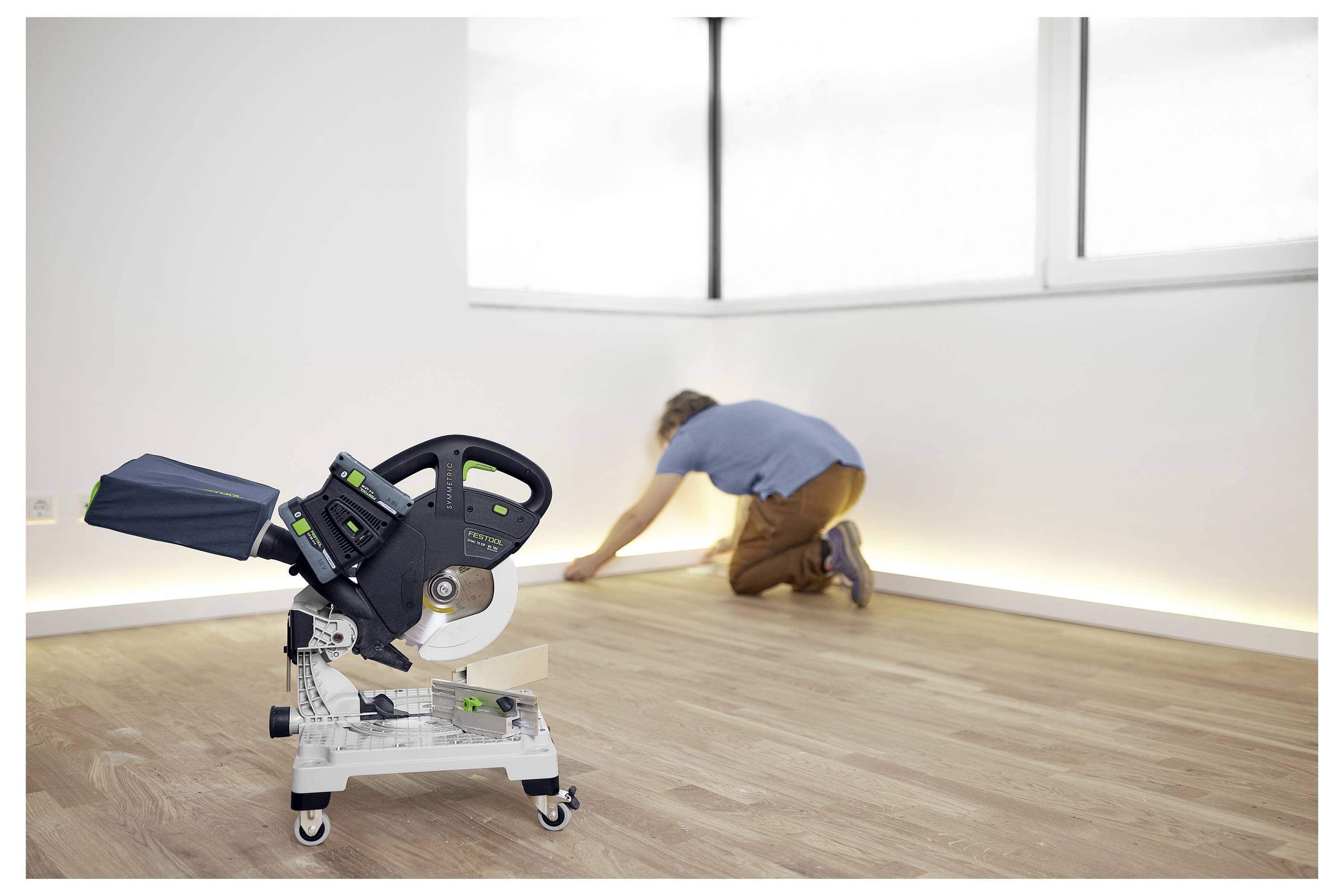 Eine Elektrosäge steht im Vordergrund auf einem Holzboden. Im Hintergrund kniet eine Person und installiert Fußleisten in einem hell beleuchteten Raum.