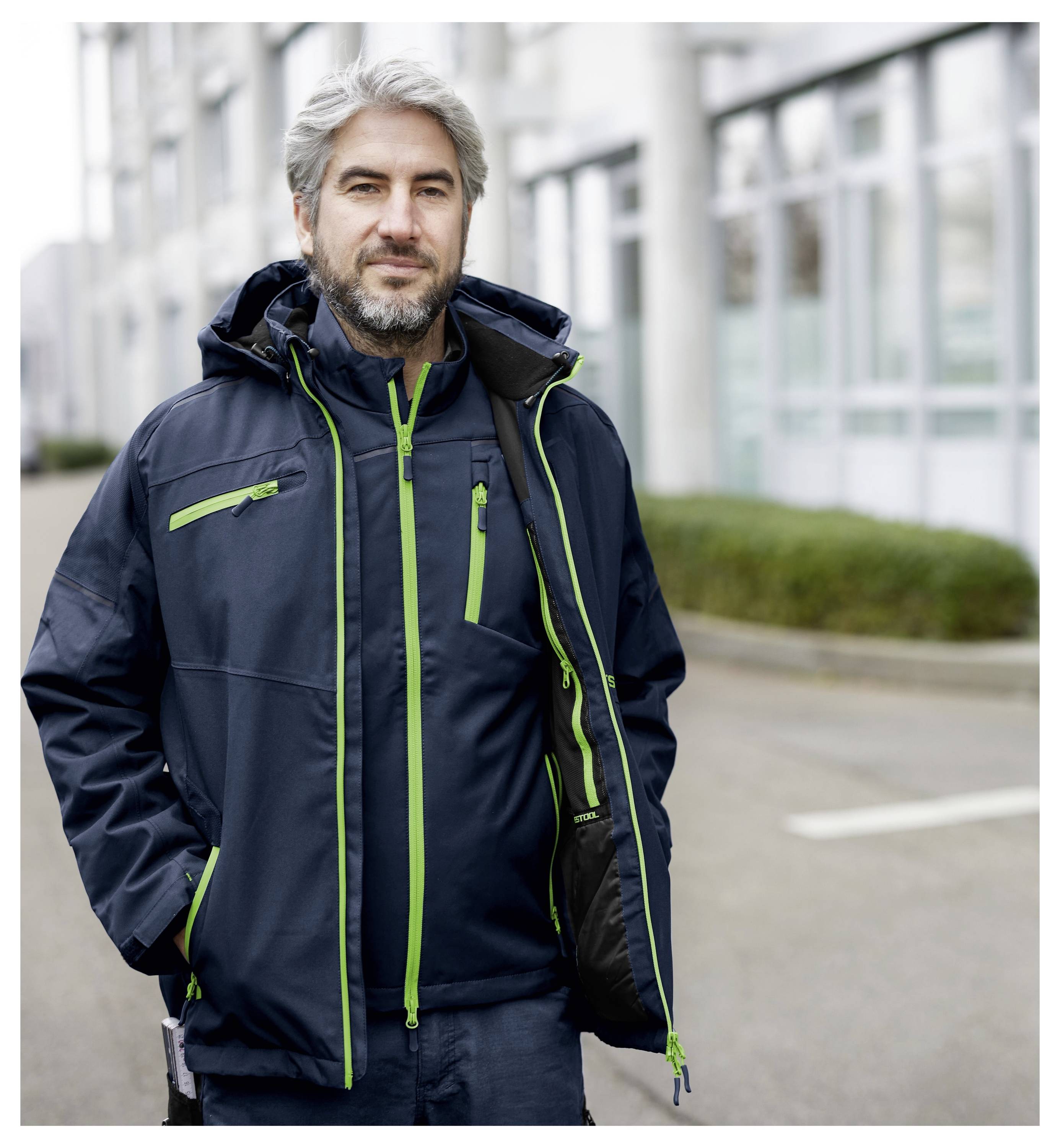 Ein Mann mit grauem Haar und Bart, der eine dunkle Jacke mit grünen Akzenten trägt, steht auf einer Straße in einem städtischen Gebiet und blickt in die Kamera.