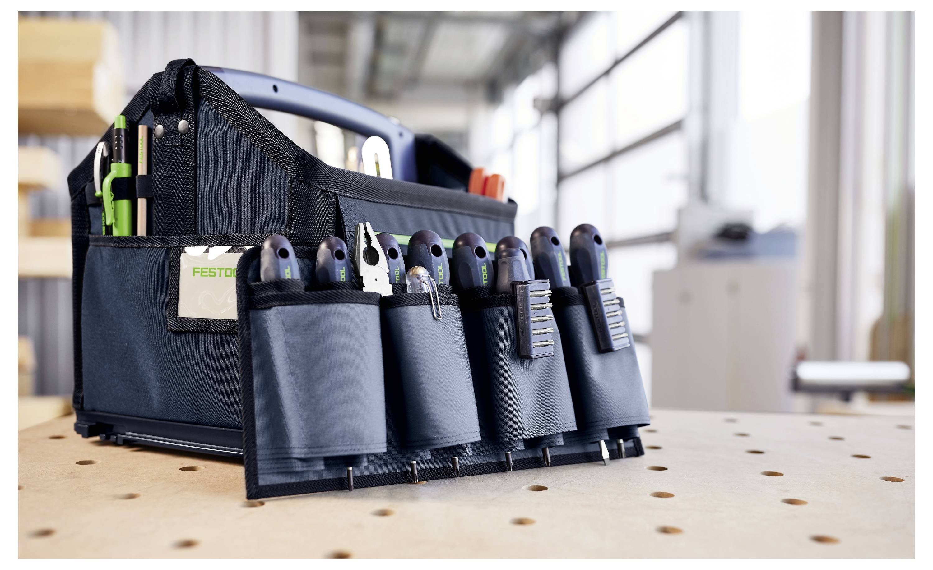Eine schwarze Werkzeugtasche mit mehreren Taschen hält verschiedene Werkzeuge wie Zangen und Schraubendreher auf einer Werkbank in einer gut beleuchteten Werkstatt.