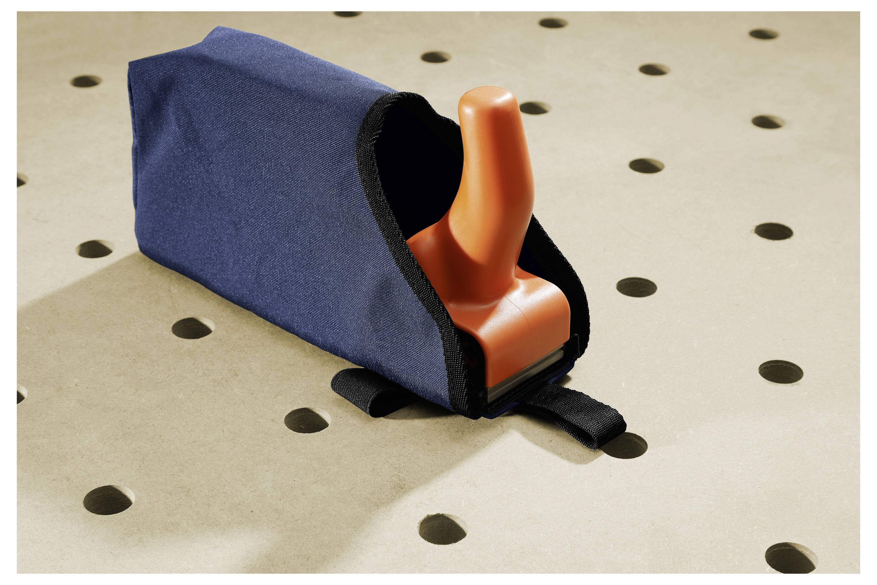 Oranger Griff, der aus einem blauem stoffbezogenem Objekt herausragt, das auf einer perforierten beigen Oberfläche ruht.