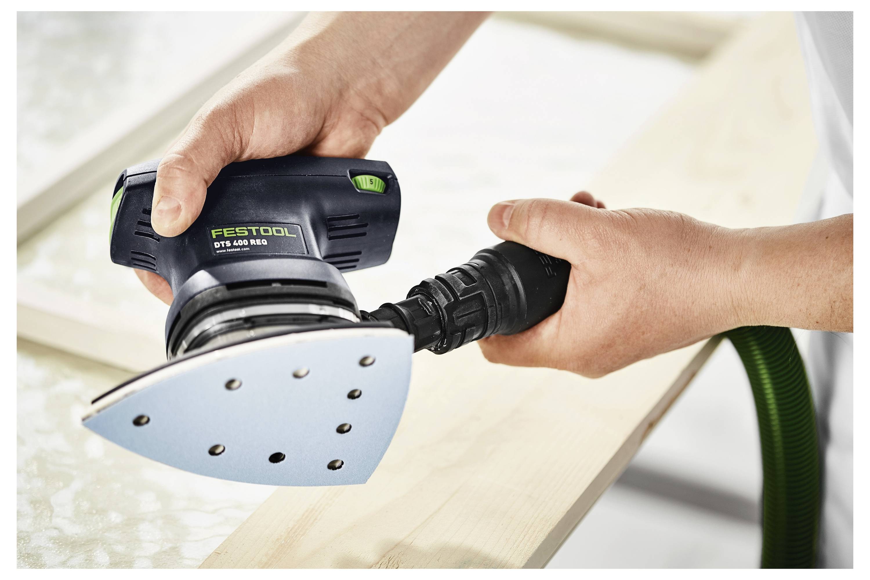 Hände, die einen Festool DTS 400 Schleifer auf einer Holzoberfläche verwenden, wobei das ergonomische Design des Werkzeugs und die Effizienz beim Glätten von Holz hervorgehoben werden.