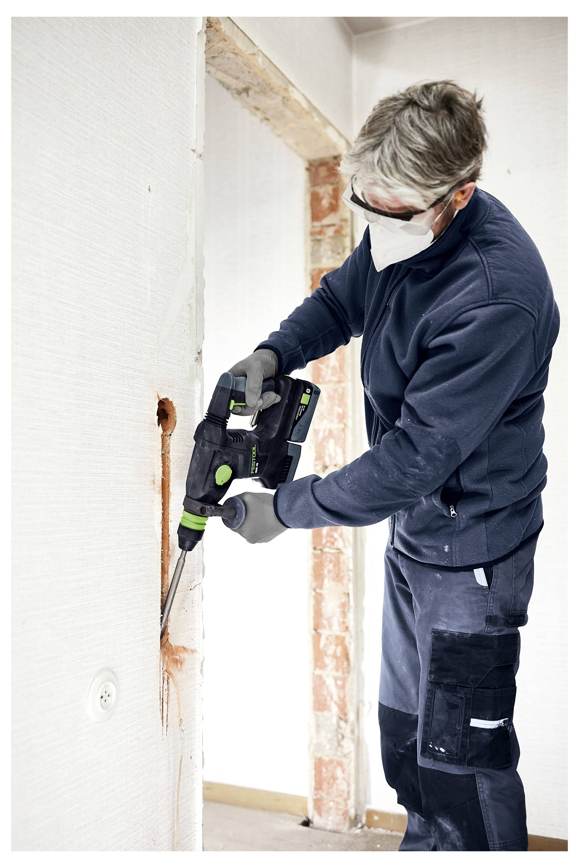 Eine Person, die ein Elektrowerkzeug benutzt, um ein Loch in eine Wand zu bohren, trägt dabei eine Schutzbrille und eine Maske, was auf eine Heimrenovierungsszene hindeutet.