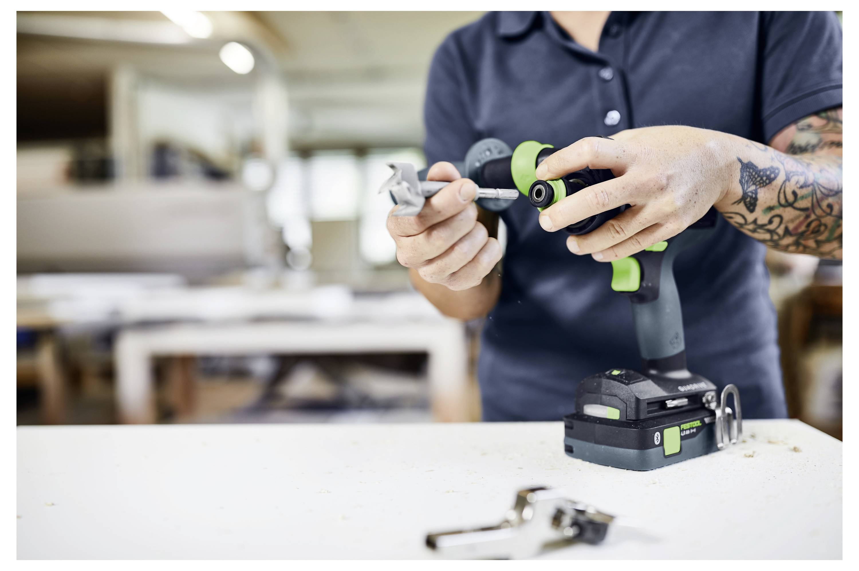 Eine Person in einer Werkstatt montiert ein Bauteil mit einem Akkubohrer. Der Raum ist mit Werkzeugen und Werkbänken ausgestattet.