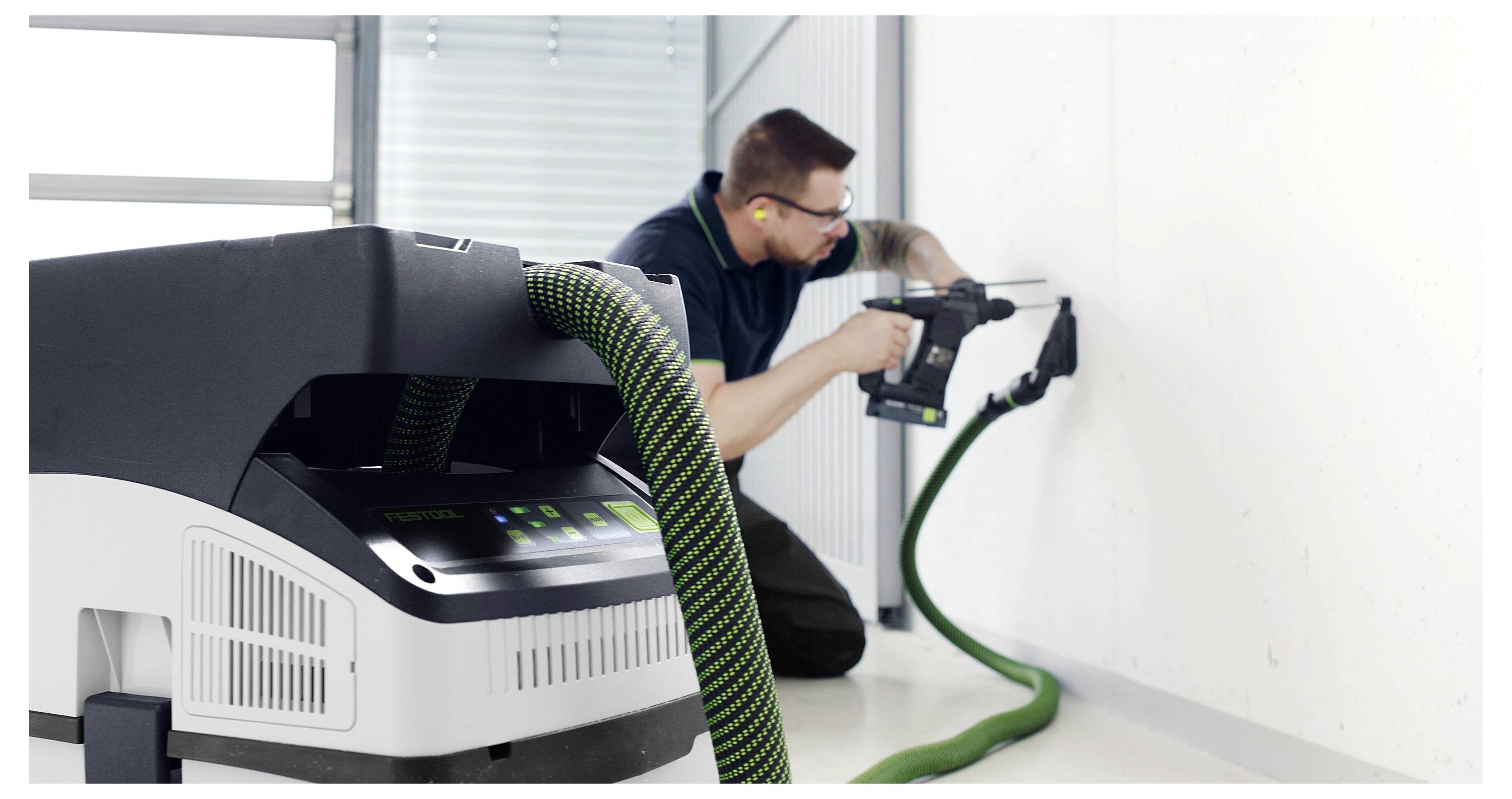 Eine Person verwendet ein Elektrowerkzeug, das mit einem Staubsauger verbunden ist, und bohrt in eine weiße Wand.