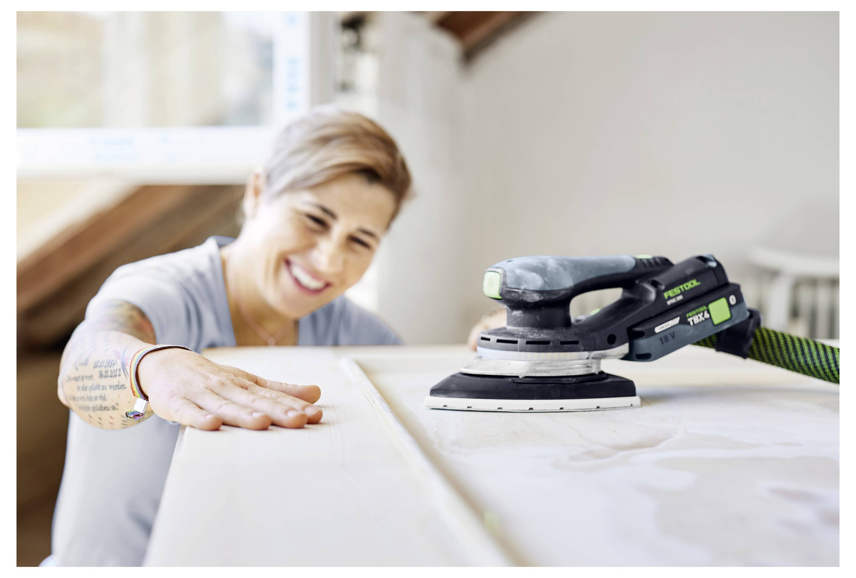 Eine lächelnde Person schleifte eine Holzoberfläche mit einem tragbaren elektischen Schleifers in einer hellen Werkstatt.