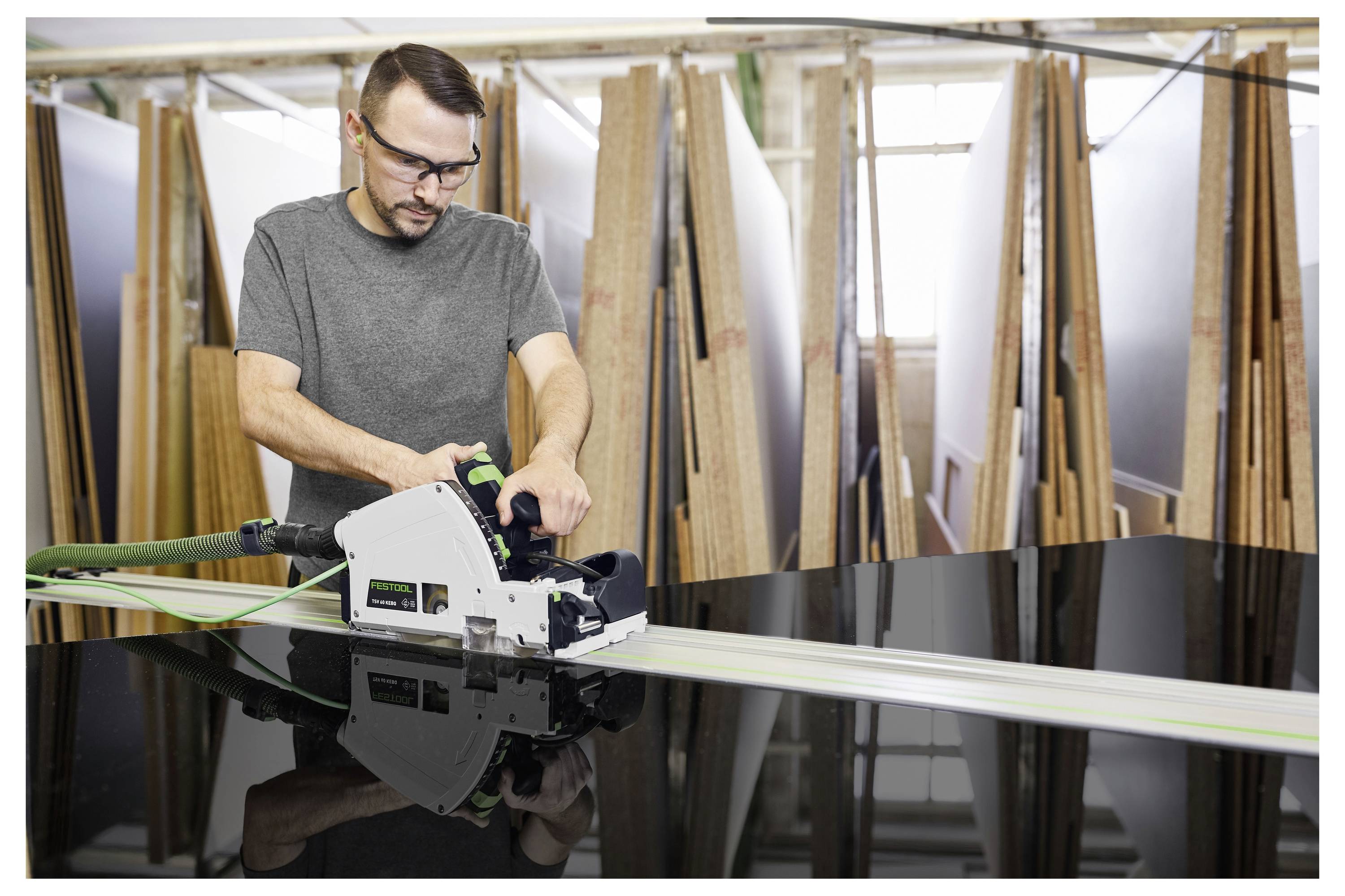 Eine Person benutzt eine Kreissäge, um ein großes schwarzes Paneel in einer Holzwerkstatt zu schneiden, umgeben von Holzbrettern und trägt eine Schutzbrille.