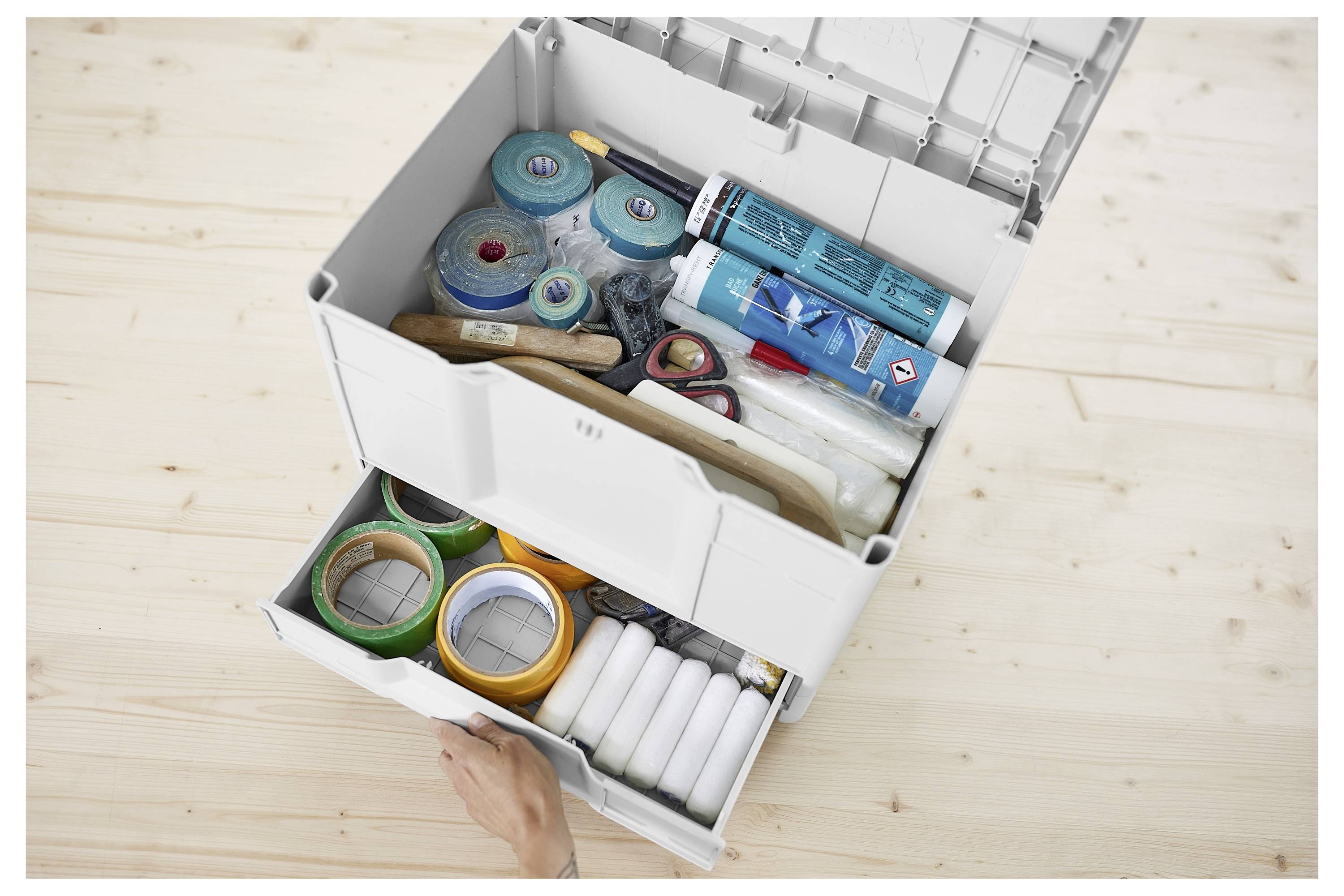 Eine Kunststoff-Aufbewahrungsbox mit zwei offenen Schubladen, die verschiedene Klebebandrollen, Röhren enthält, und eine Hand zieht die untere Schublade auf einem Holzboden.
