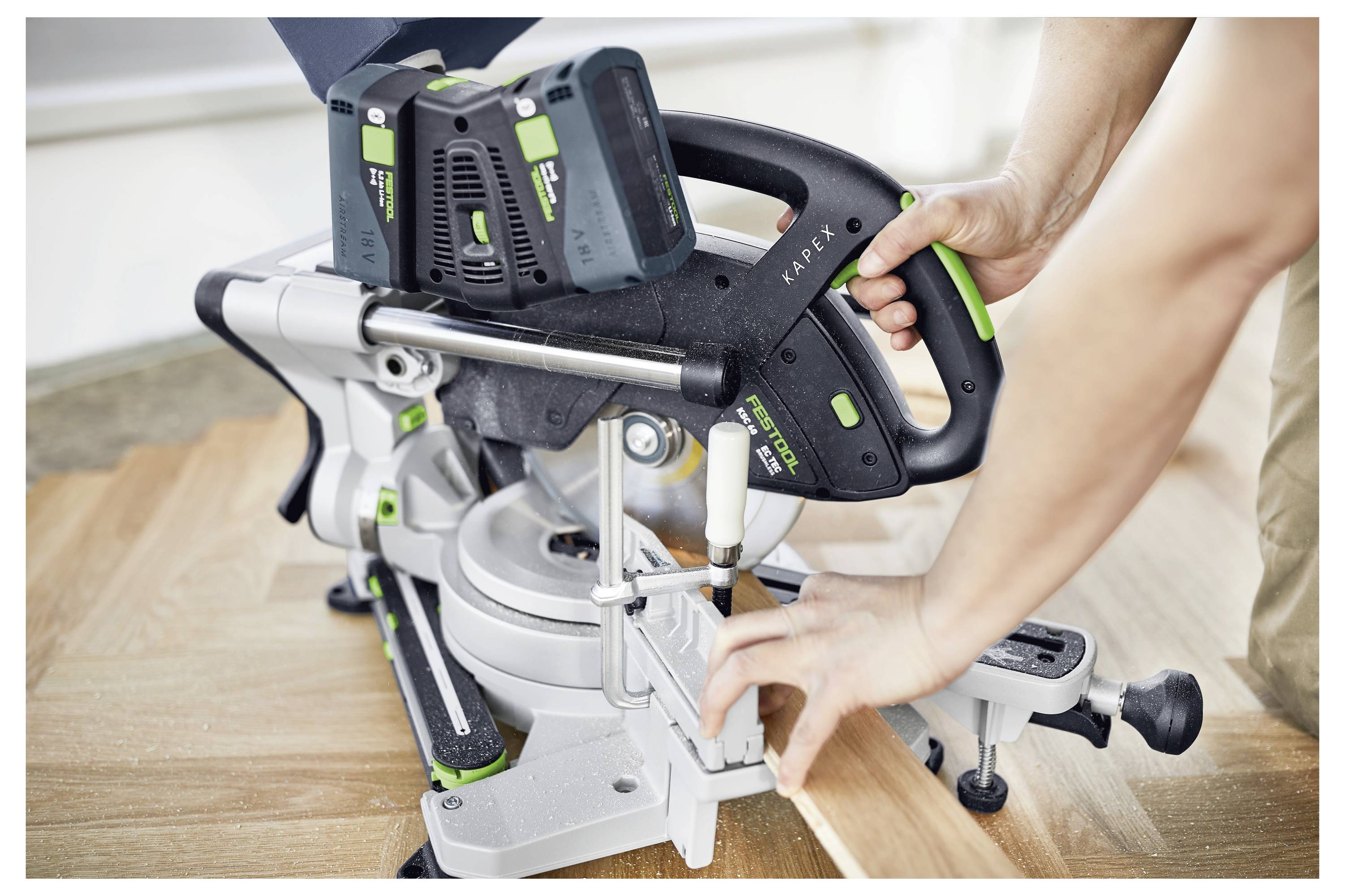 Eine Person, die Schutzbrille trägt, benutzt einen Bohrer an einem Stück weißem Holz in einer Werkstatt. Schränke und andere Werkzeuge befinden sich im Hintergrund.