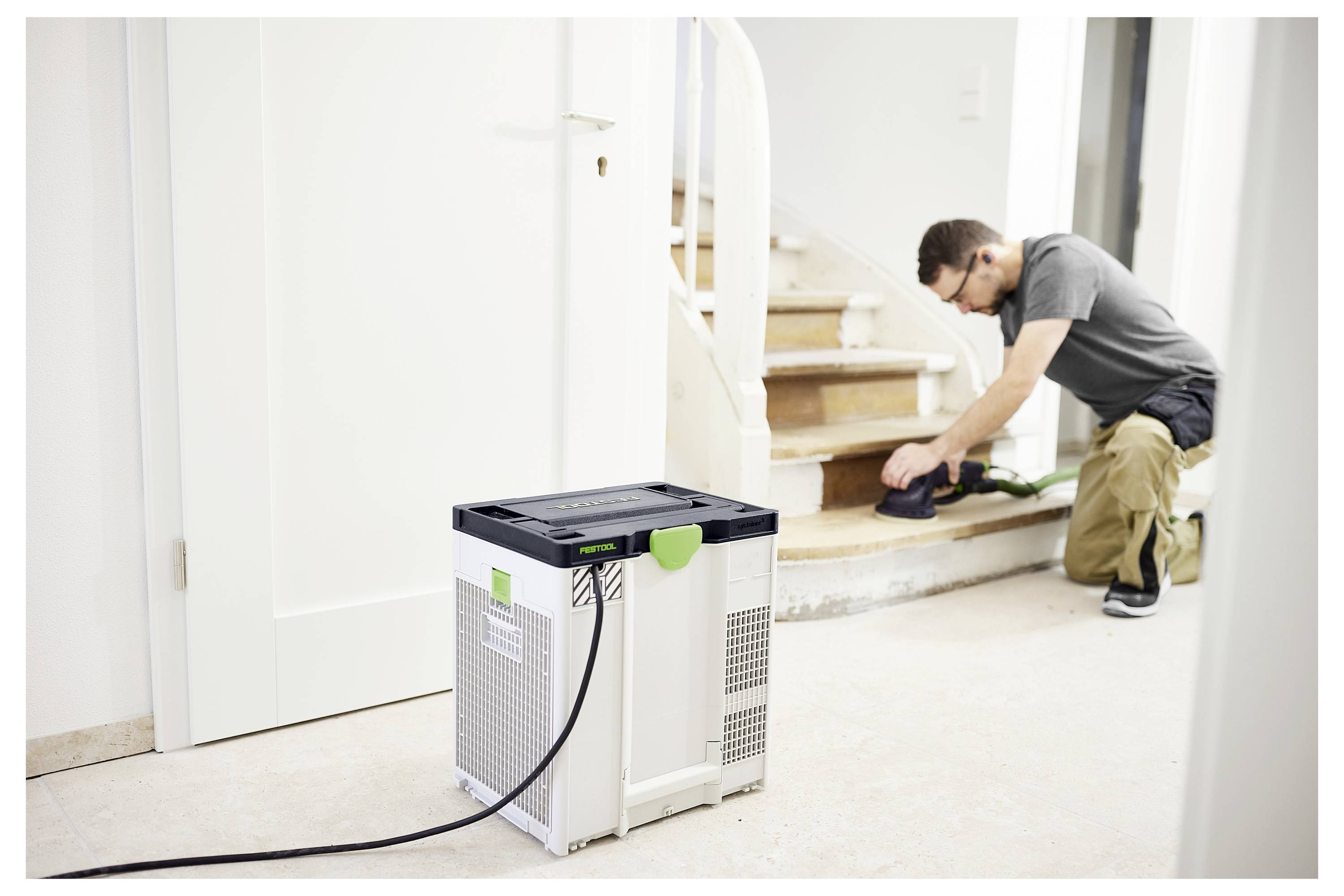 Eine Person schleift Treppen ab und trägt Gehörschutz. Ein Staubabscheider befindet sich im Vordergrund, verbunden mit dem Schleifdisketten, was die Werkstattssicherheit unterstreicht.