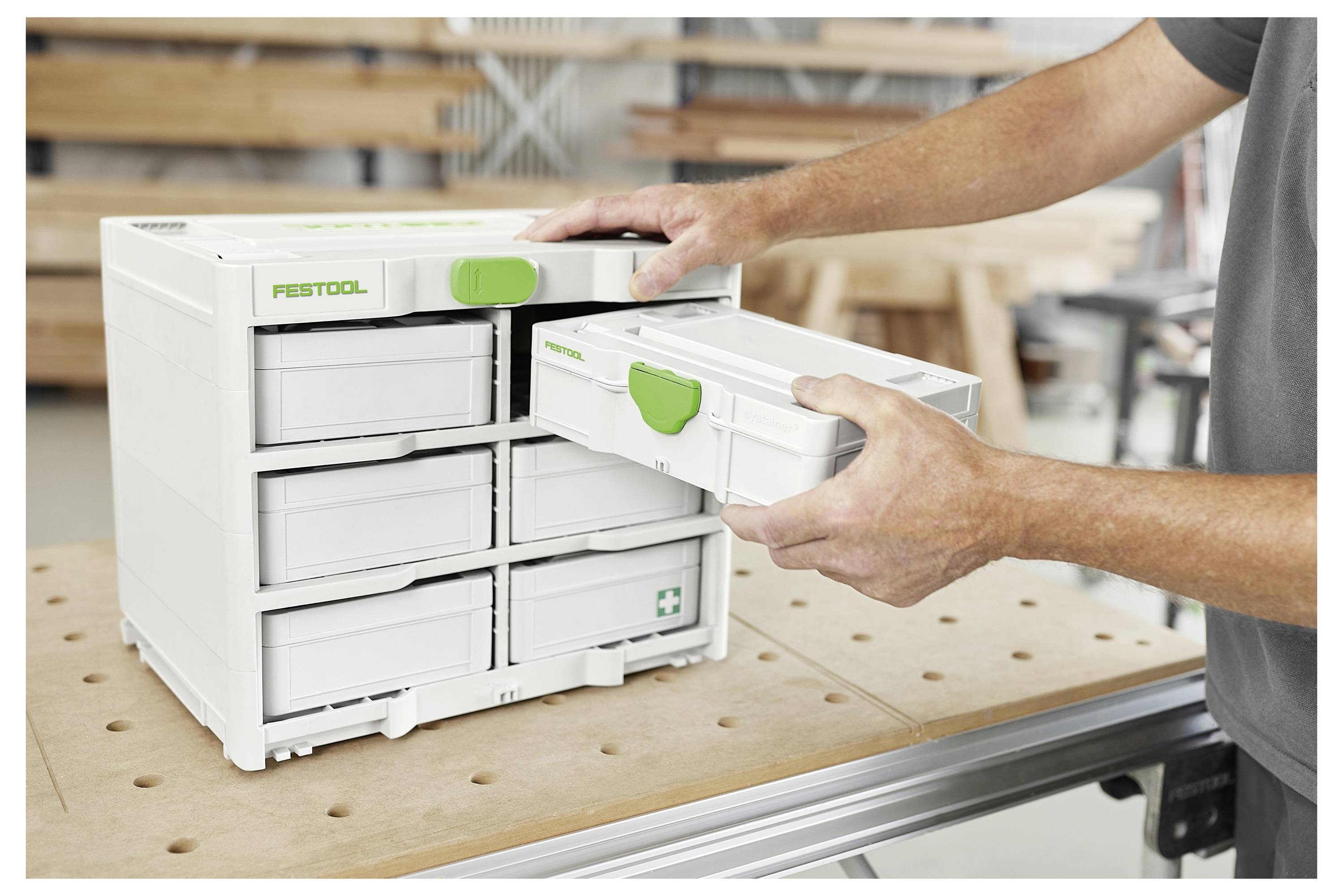 Eine Person öffnet eine Schublade eines weißen Festool-Lagerschranks auf einer Werkbank und hält einen abnehmbaren Behälter mit einem grünen Griff.