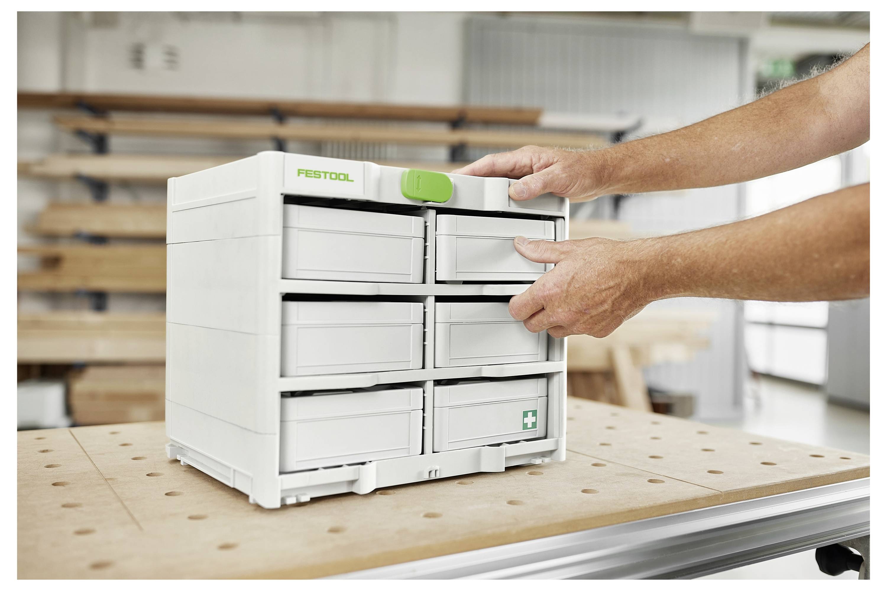 Eine Person öffnet eine sys-dock Systainer-Lagerschublade in einer Werkstatt-Umgebung, mit Holzbearbeitungswerkzeugen und Materialien, die im Hintergrund sichtbar sind.