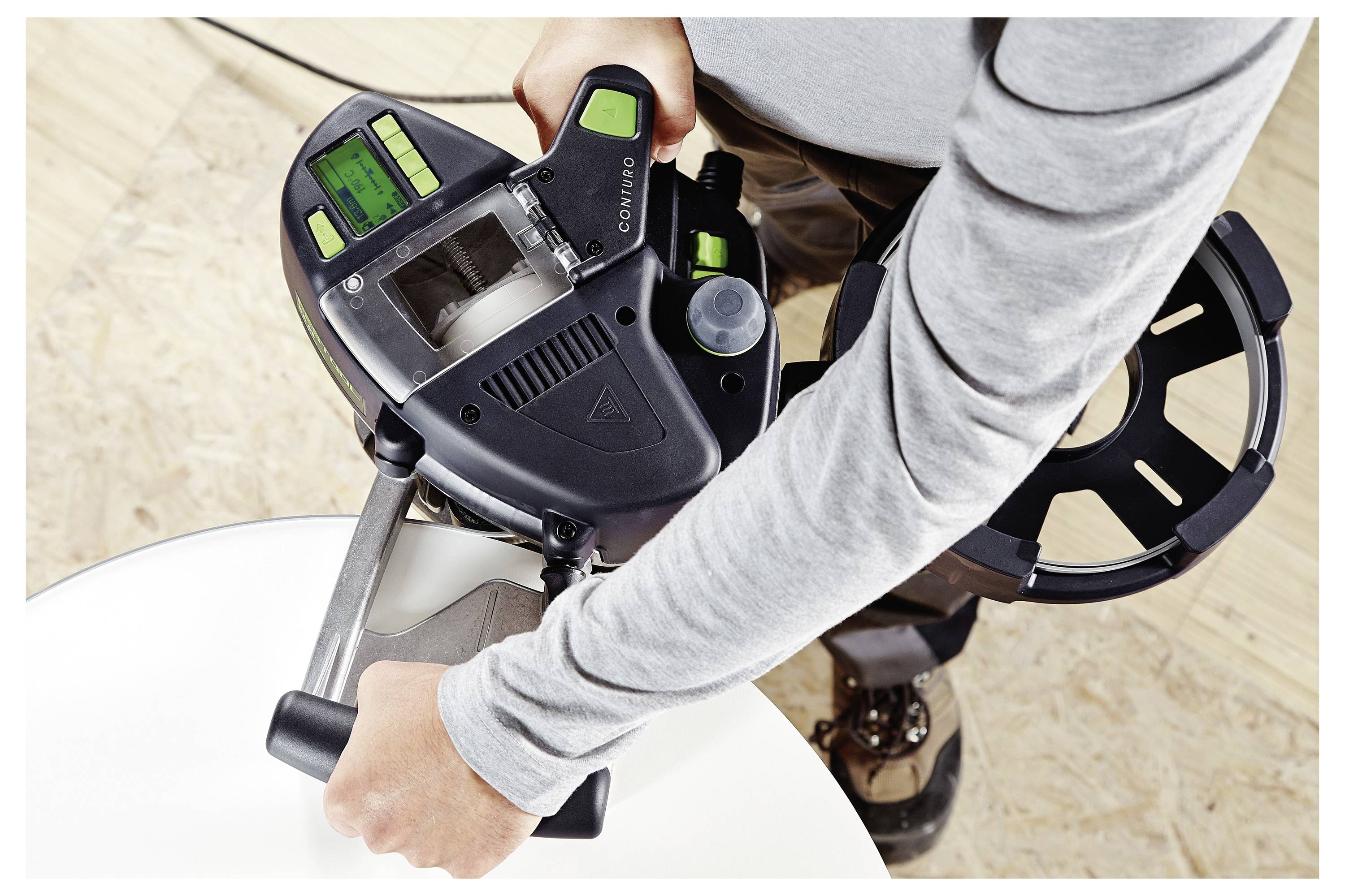 Eine Person benutzt ein Elektrowerkzeug mit digitaler Anzeige, um an einer Holzoberfläche zu arbeiten. Das Werkzeug ist durch zwei Griffe gesichert und über dem Holz positioniert.