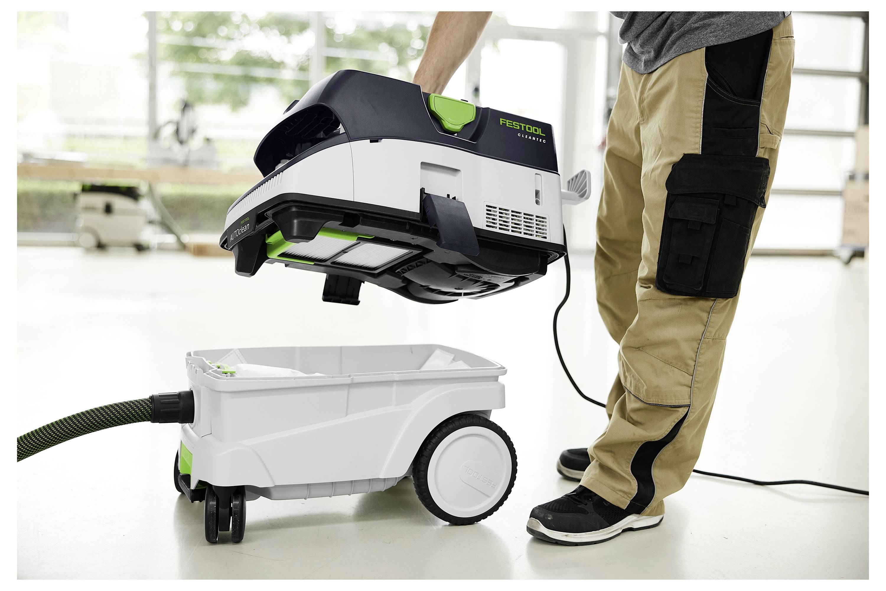 Eine Person montiert einen weiß-schwarzen Festool-Staubsauger, steht auf einem hellen Boden, trägt eine beige Hose und schwarze Sneaker.