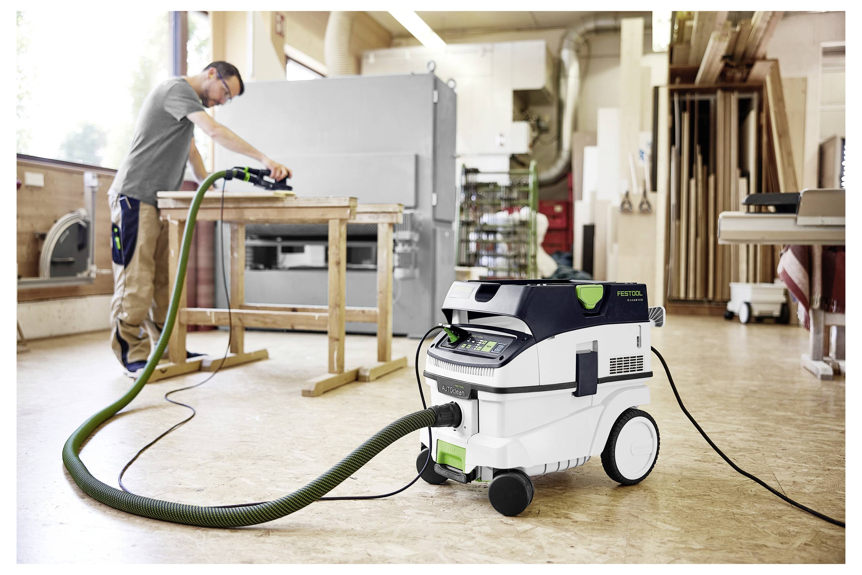 Eine Person benutzt ein Elektrowerkzeug, das mit einem Festool-Staubsauger in einer Werkstatt verbunden ist, umgeben von Holzbearbeitungsgeräten und Materialien.