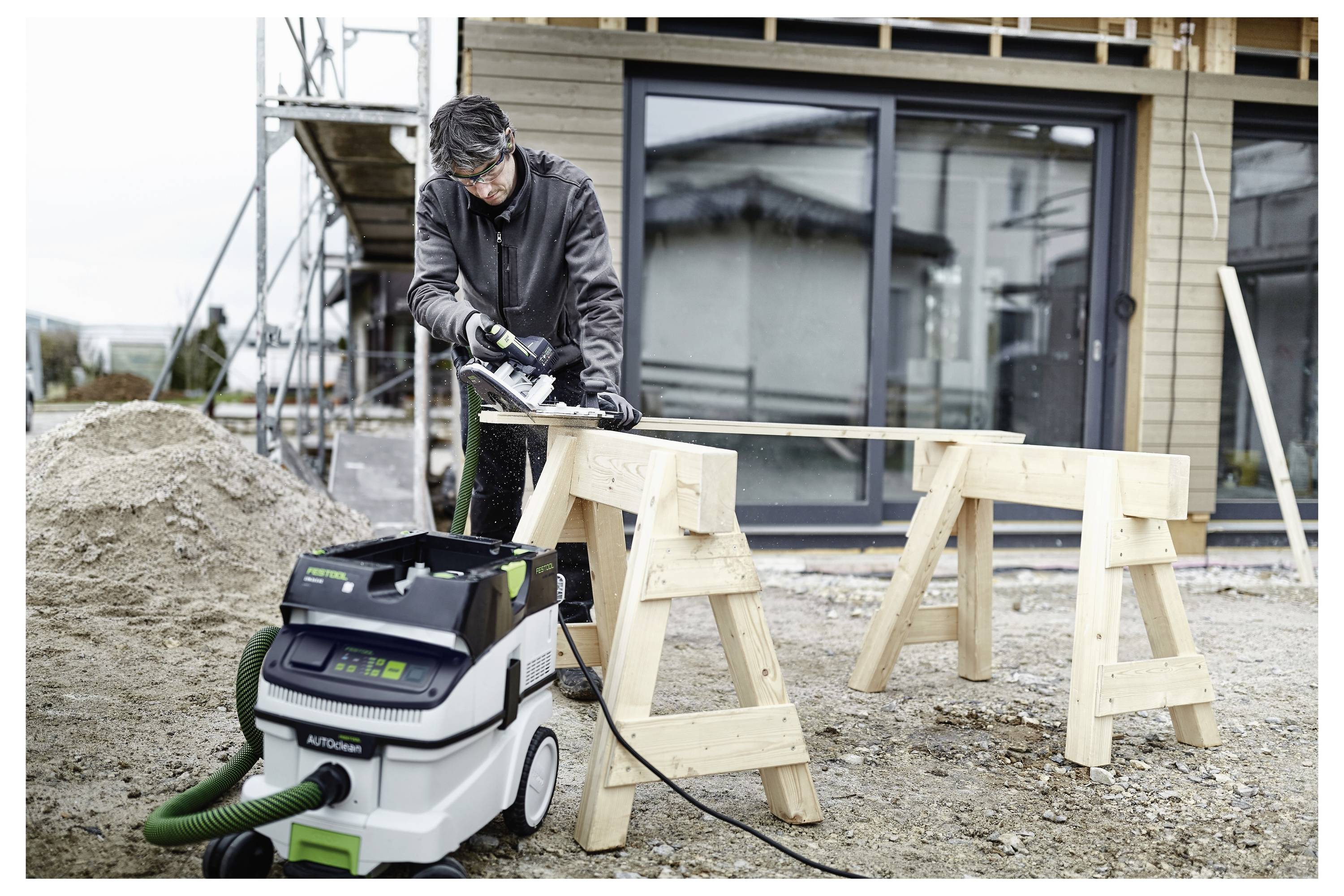 Eine Person in Schutzausrüstung benutzt eine elektrische Säge auf einem Holzbrett, das von Sägeböcken gestützt wird, außerhalb eines modernen Gebäudes, das sich im Bau befindet.