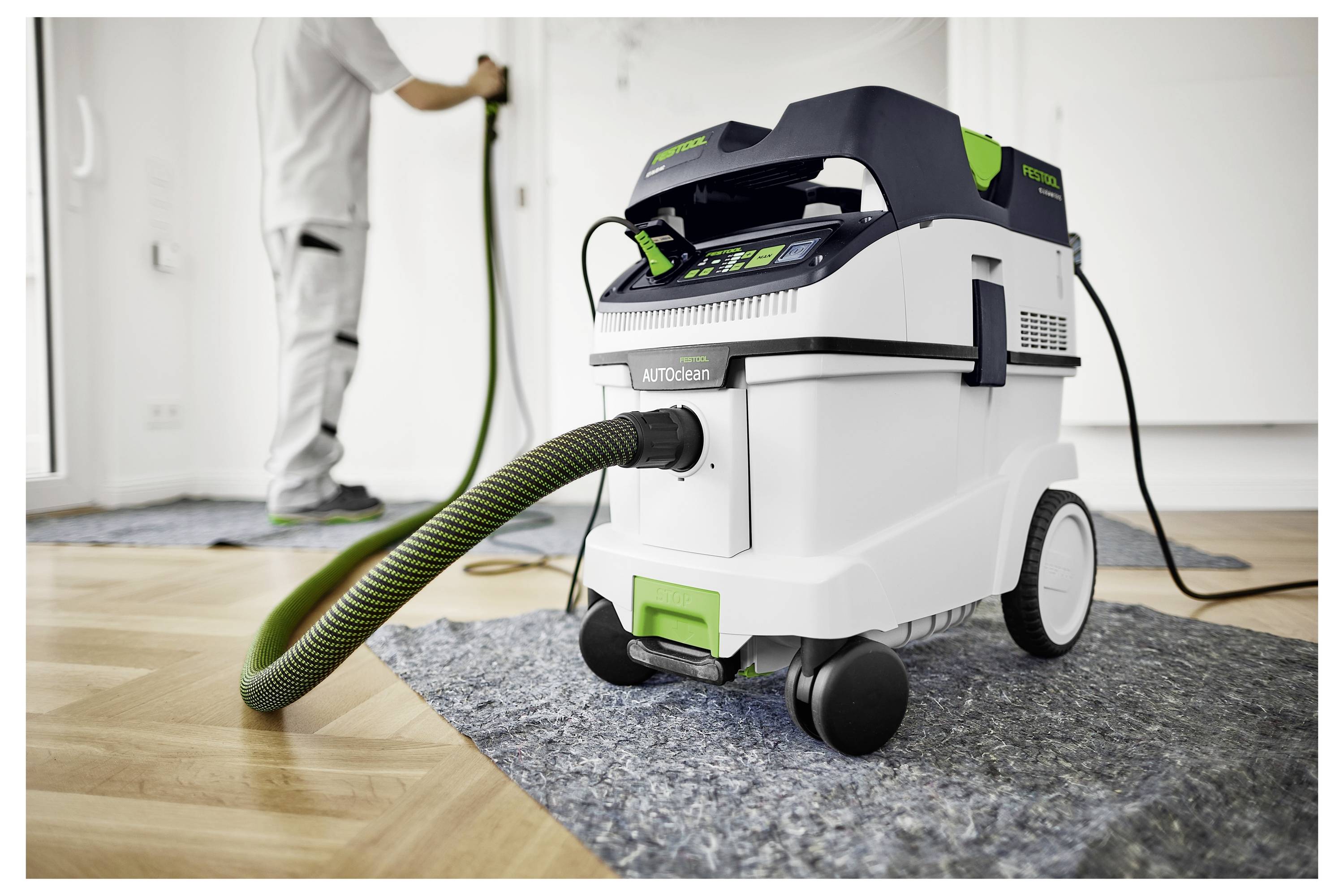 „Ein Staubsauger mit Schlauch steht auf einem Holzboden und konzentriert sich auf eine Reinigungsaufgabe. Eine Person im Hintergrund benutzt den Staubsauger.