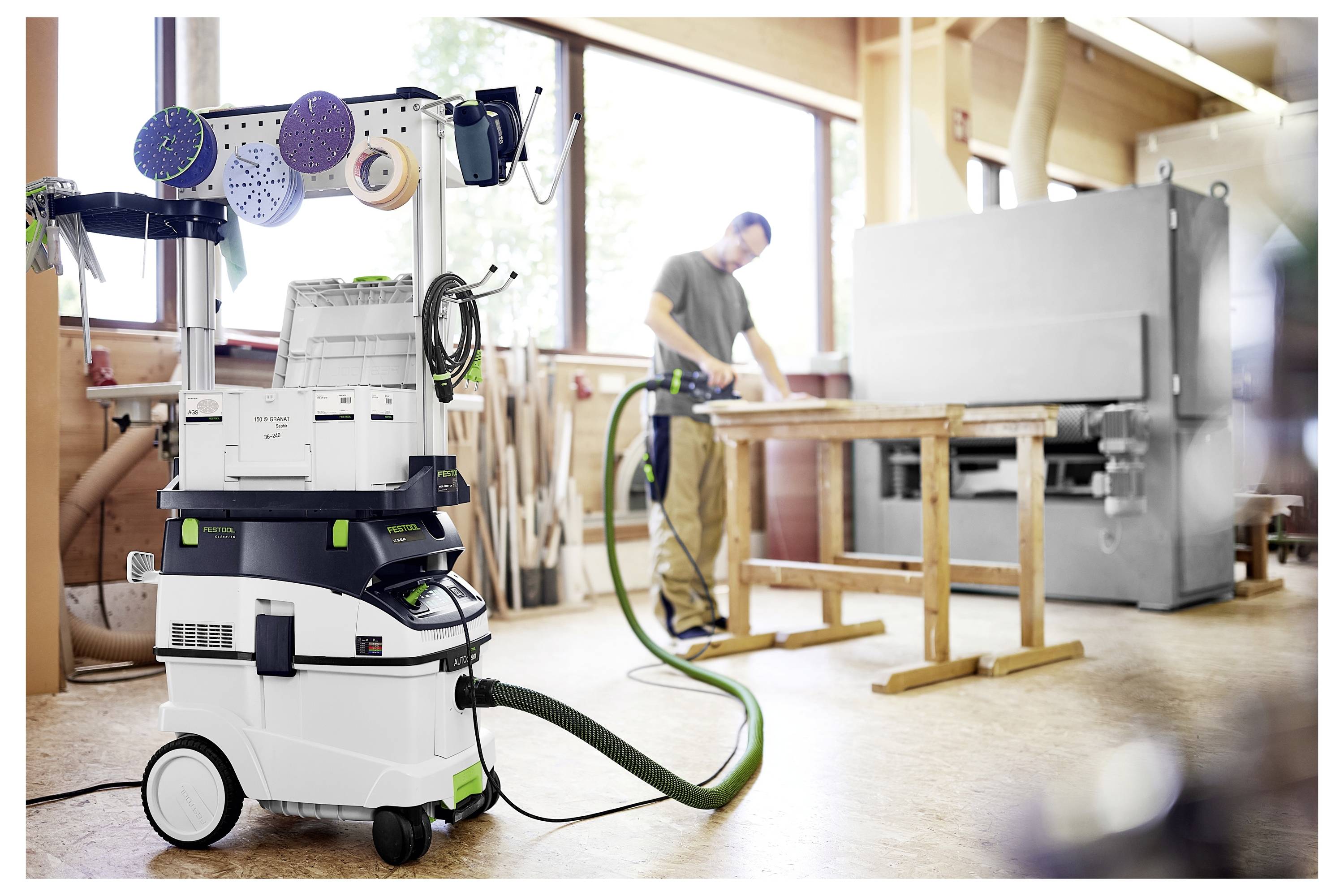 Ein Holzarbeiter benutzt eine Schleifmaschine in einer Werkstatt; ein Staubabscheider steht im Vordergrund und ist mit der Maschine durch einen Schlauch verbunden.
