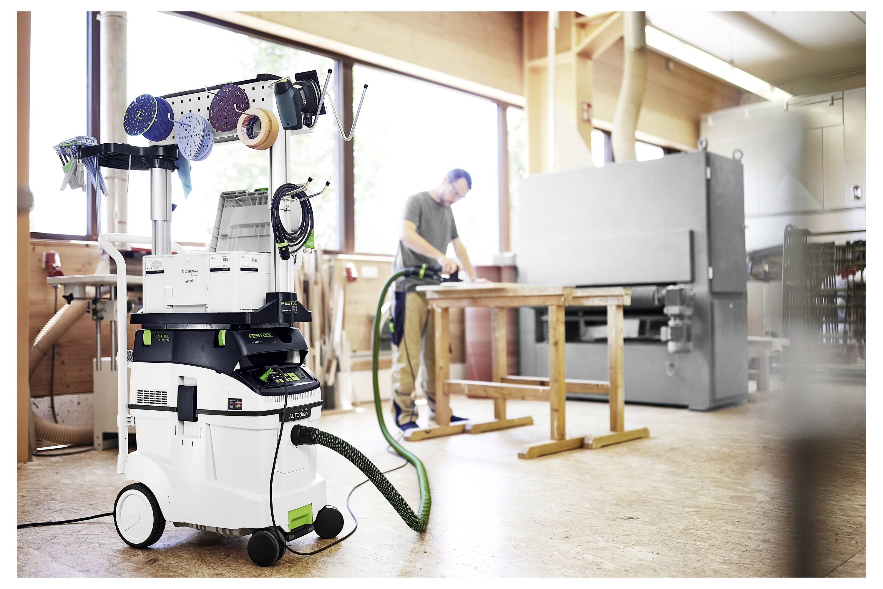 Ein Werkstatt mit einer Person, die Holzbearbeitungswerkzeuge benutzt, ein Staubabsauger mit Werkzeugen im Vordergrund und ein Fenster mit natürlichem Licht.