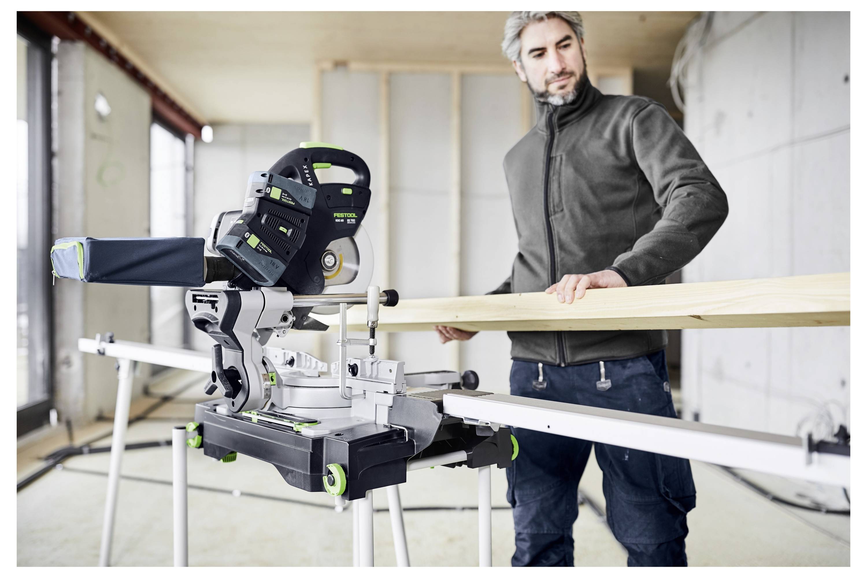 Eine Person in einer Werkstatt benutzt eine Gehrungssäge, um ein Holzbrett zu schneiden. Die Umgebung ist ein teilweise konstruierter Raum mit unvollendeten Wänden.