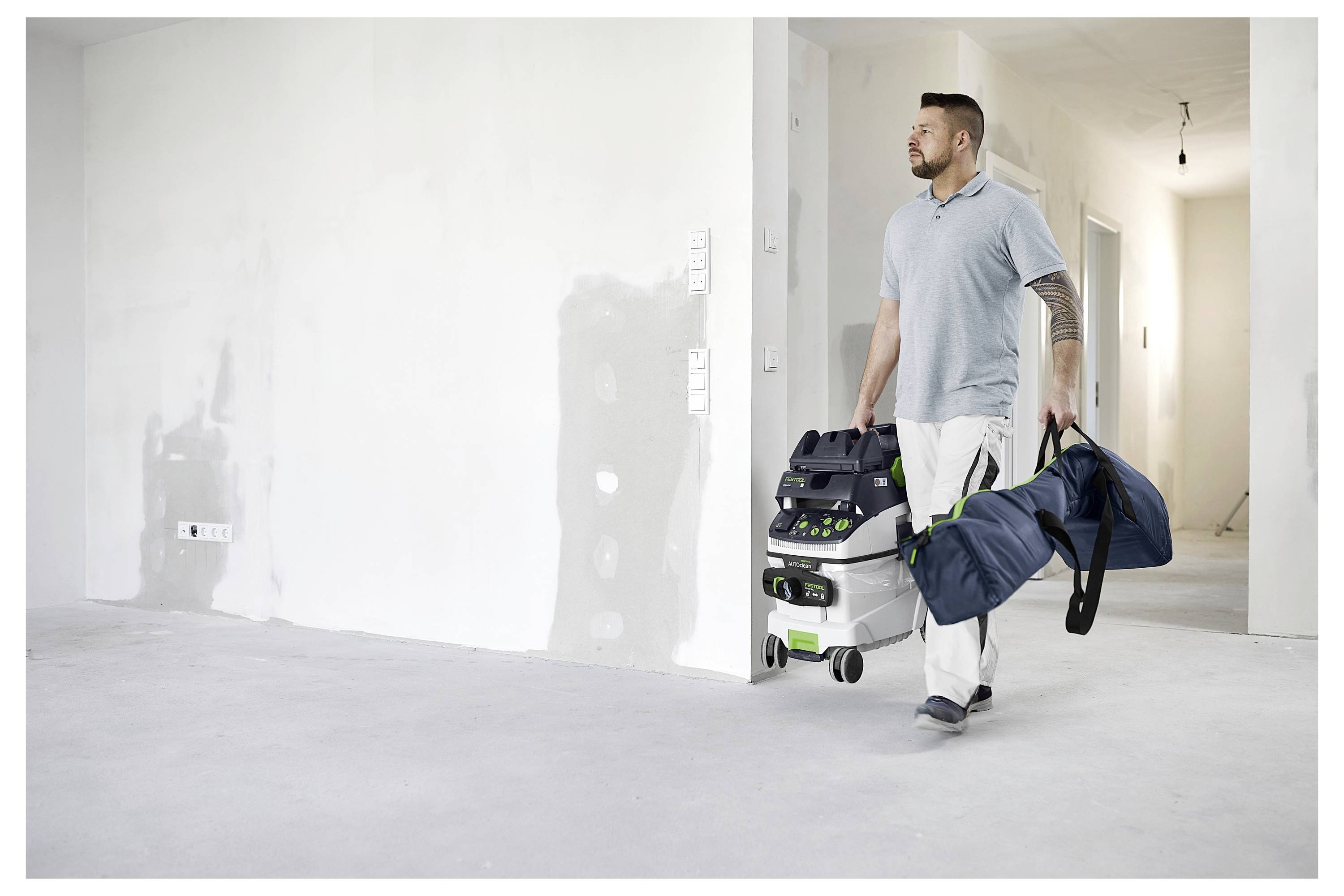 Eine Person in Arbeitskleidung trägt einen tragbaren Staubsauger und eine Werkzeugtasche durch einen unvollendeten Raum mit Flicken an den Wänden.