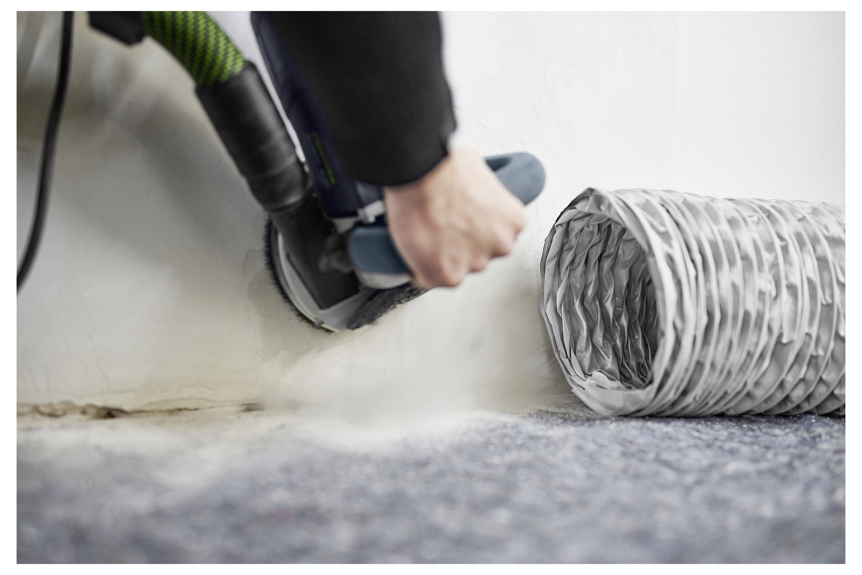 Person in Schutzausrüstung reinigt Schimmel aus einer weißen Wandecke, wobei spezialisierte Geräte auf einem kunststoffabgedeckten Boden verwendet werden.