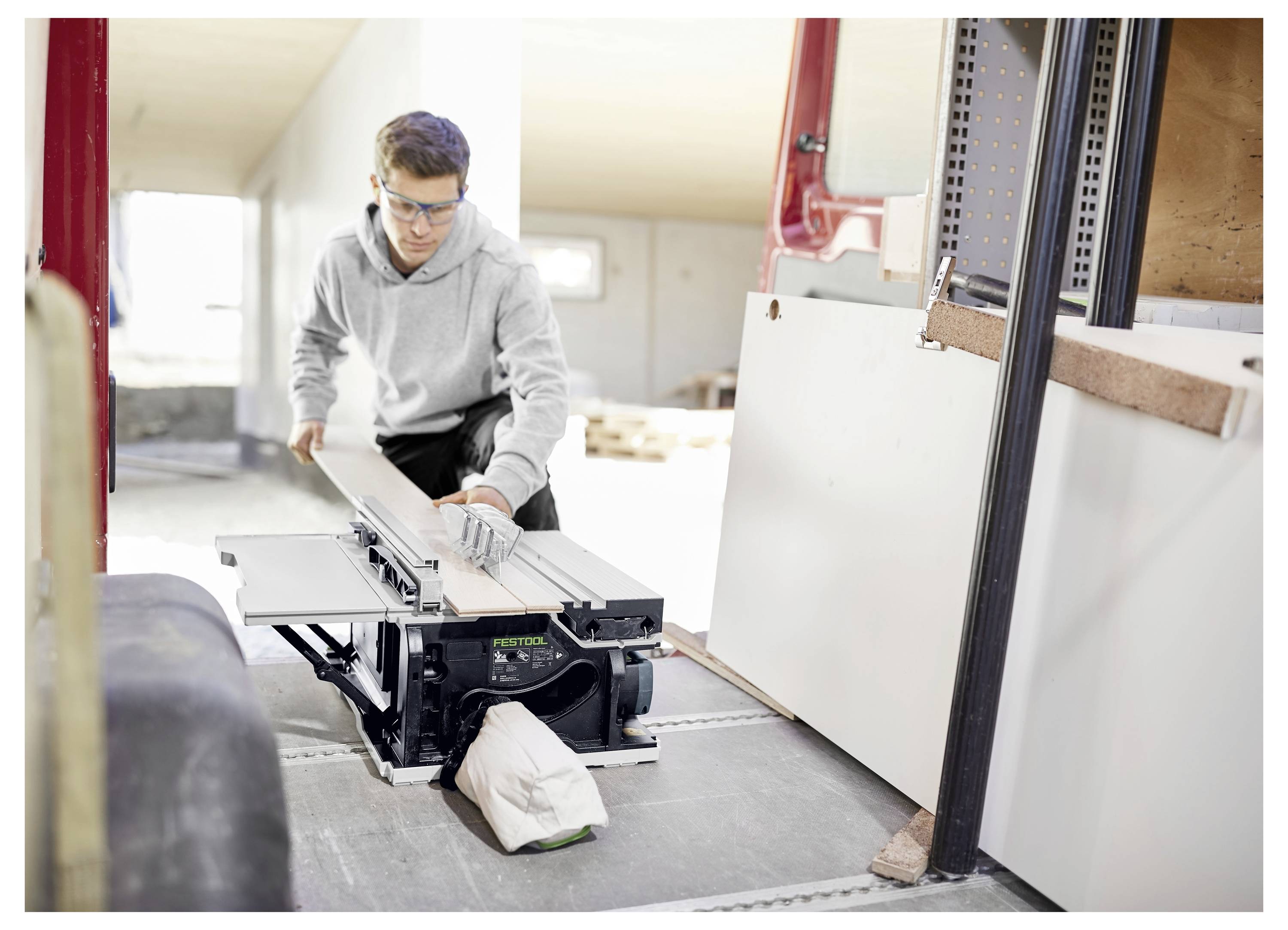 Person using a table saw to cut wood inside a workshop. They are wearing protective eyewear and a gray hoodie, focused on their task.