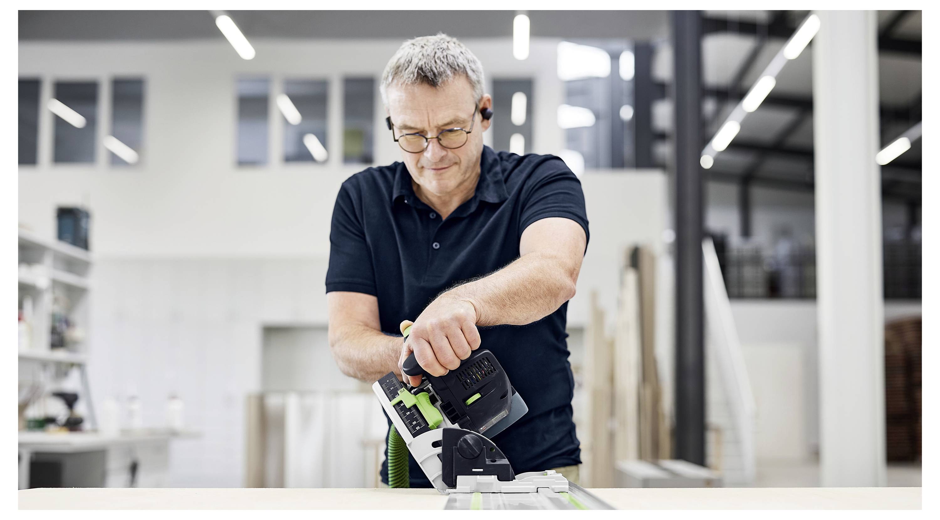 Ein Mann benutzt ein Werkzeug in einer Werkstatt. Er trägt eine Brille und ein schwarzes Poloshirt und konzentriert sich auf seine Aufgabe in einem hellen, modernen Arbeitsbereich.