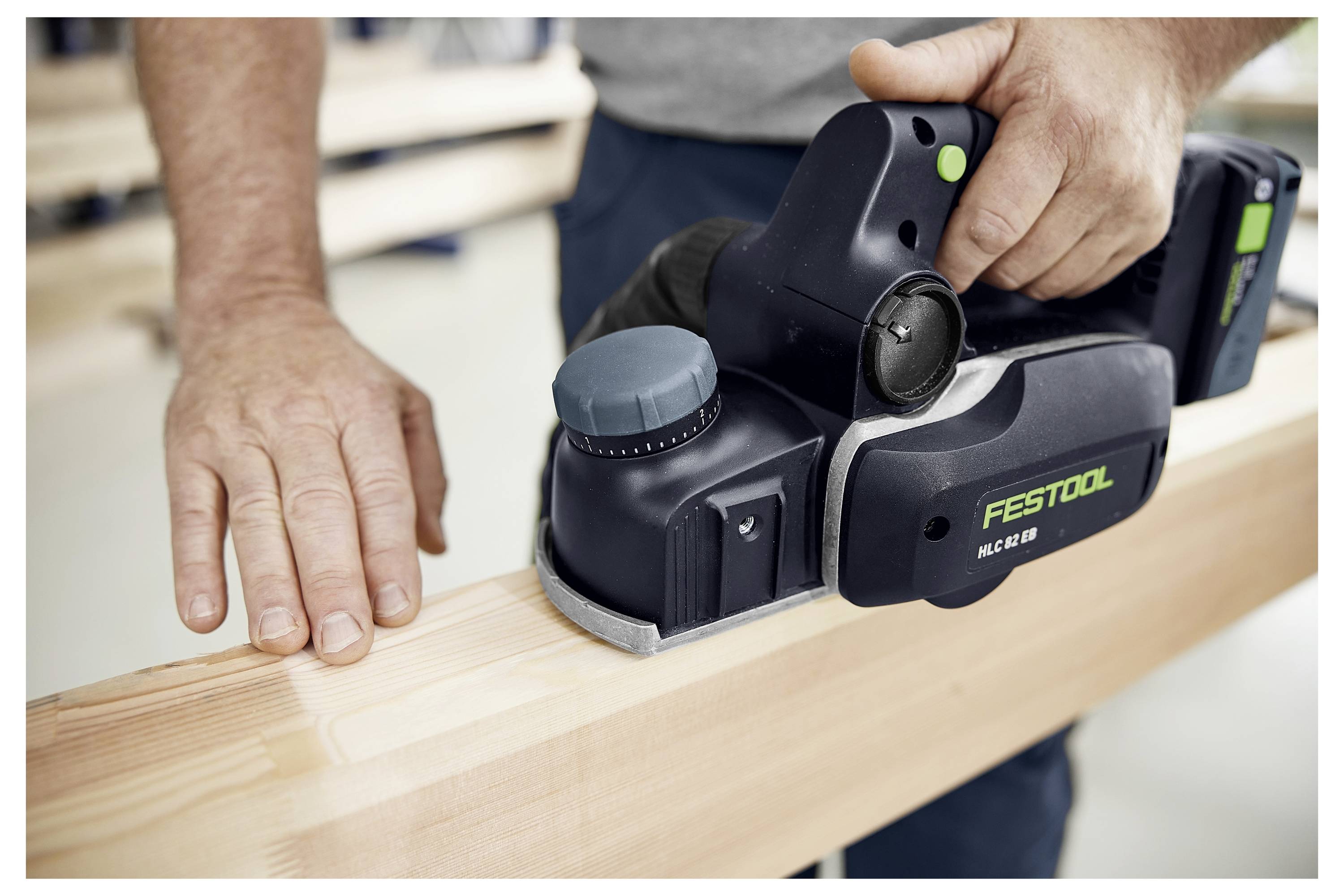 Eine Person, die einen Festool HG 82 EB Elektro-Handhobel auf einem Holzbalken verwendet. Das Werkzeug hat ein schwarz-grünes Design, wobei die Hand der Person das Holz führt.