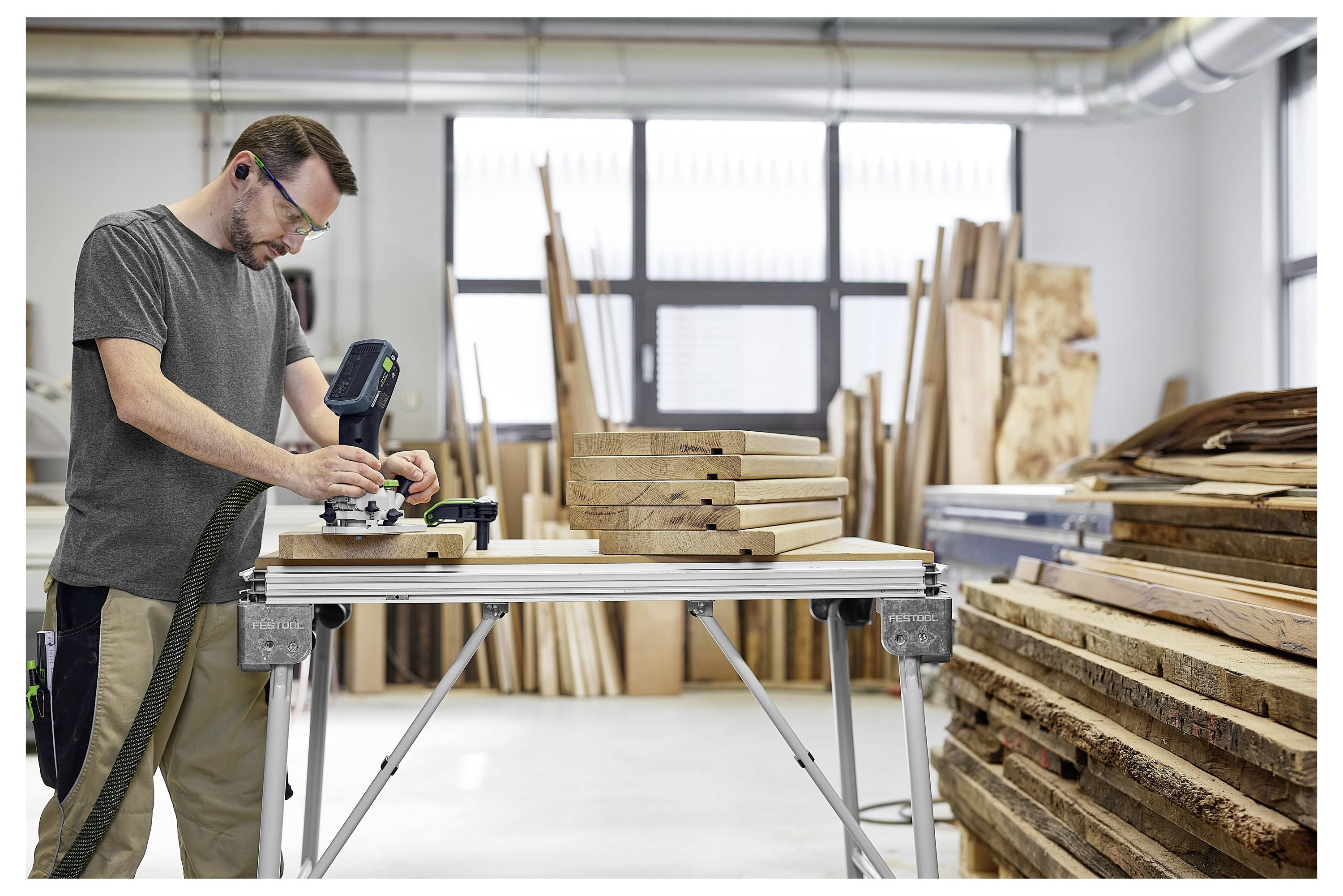 Ein Mann mit einer Schutzbrille benutzt ein Elektrowerkzeug auf Holzbrettern in einer Werkstatt, die mit Holzbrettern gefüllt ist und in die Sonnenlicht von großen Fenstern fällt.