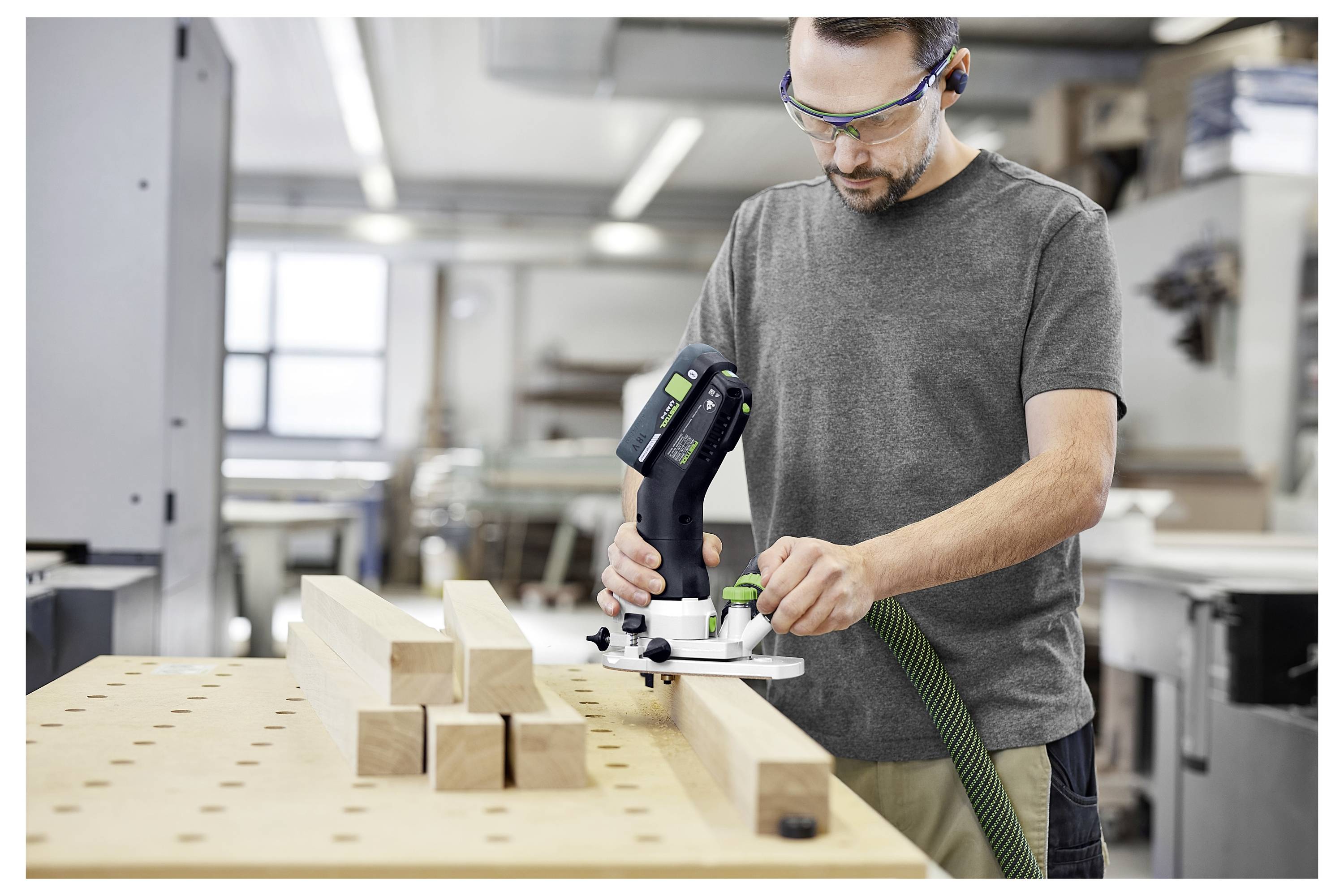 Mann, der ein Elektrowerkzeug auf Holzbalken in einer Werkstatt benutzt; trägt Schutzbrille und konzentriert sich auf die Aufgabe.